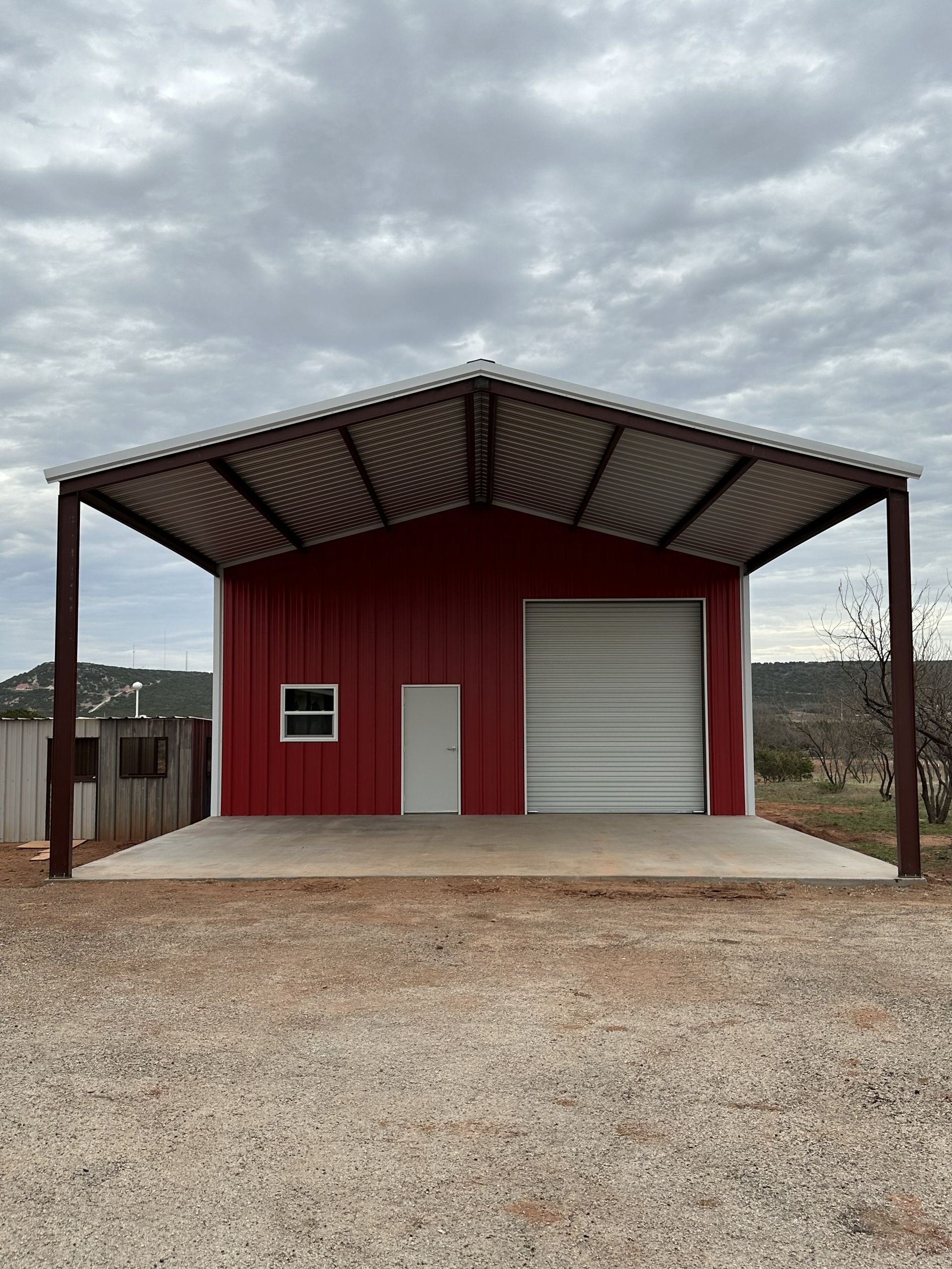 Red metal garage building with a large roll-up door and covered concrete pad under a cloudy sky