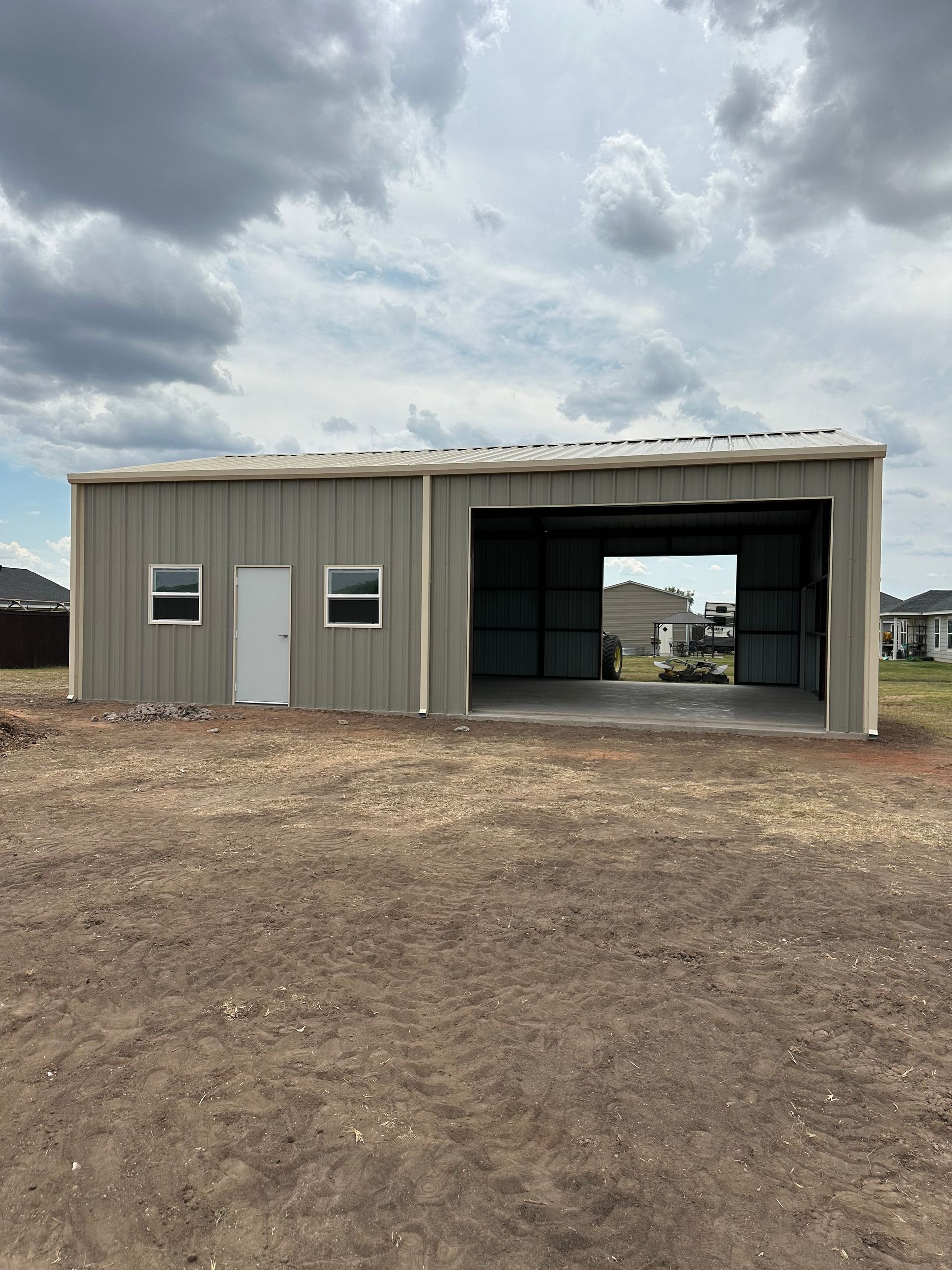 Small beige metal building with three windows and a large open garage bay on a dirt lot under cloudy skies