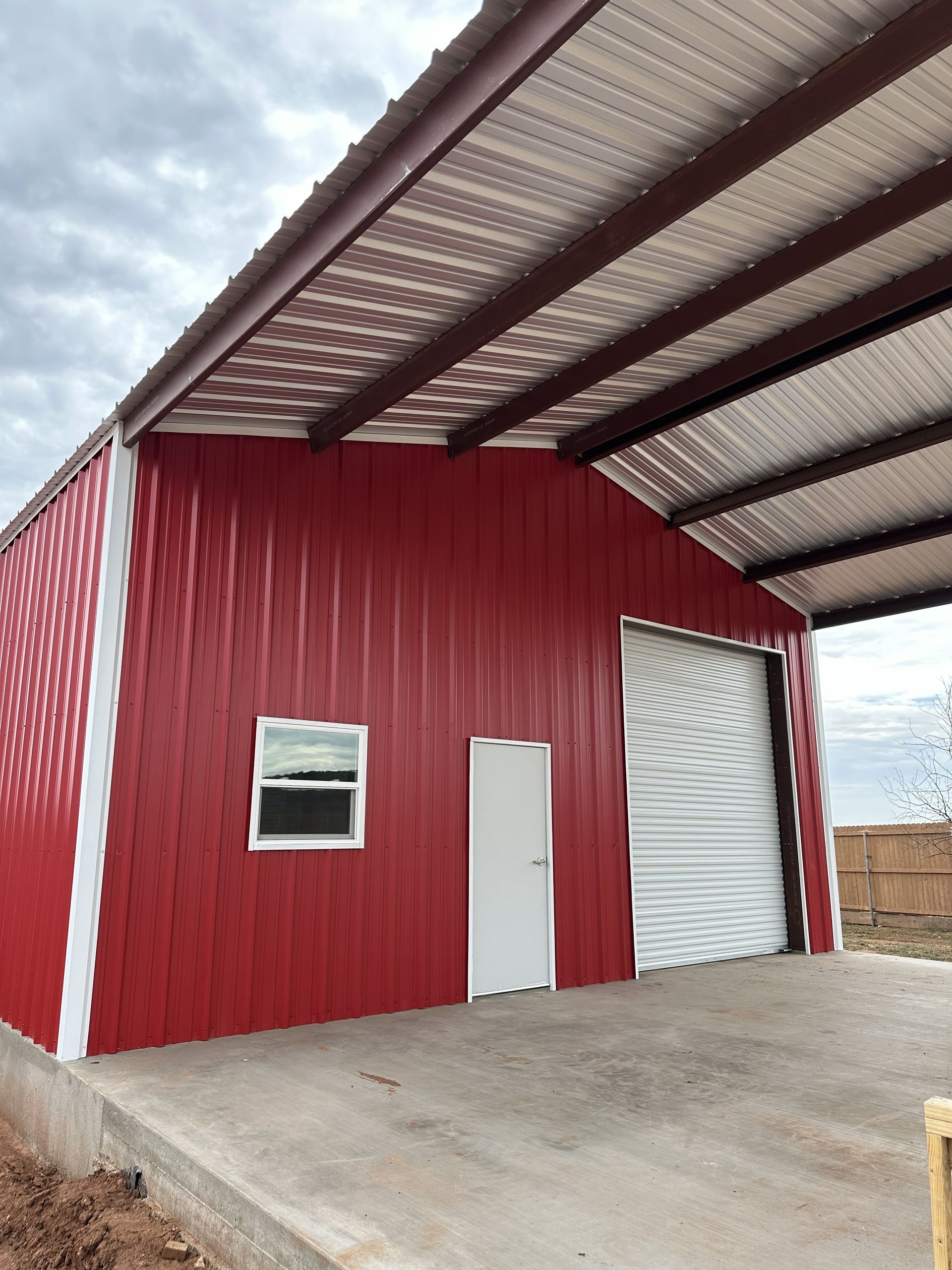 Red metal warehouse with a white door, small window, and large roll-up garage door under a canopy.