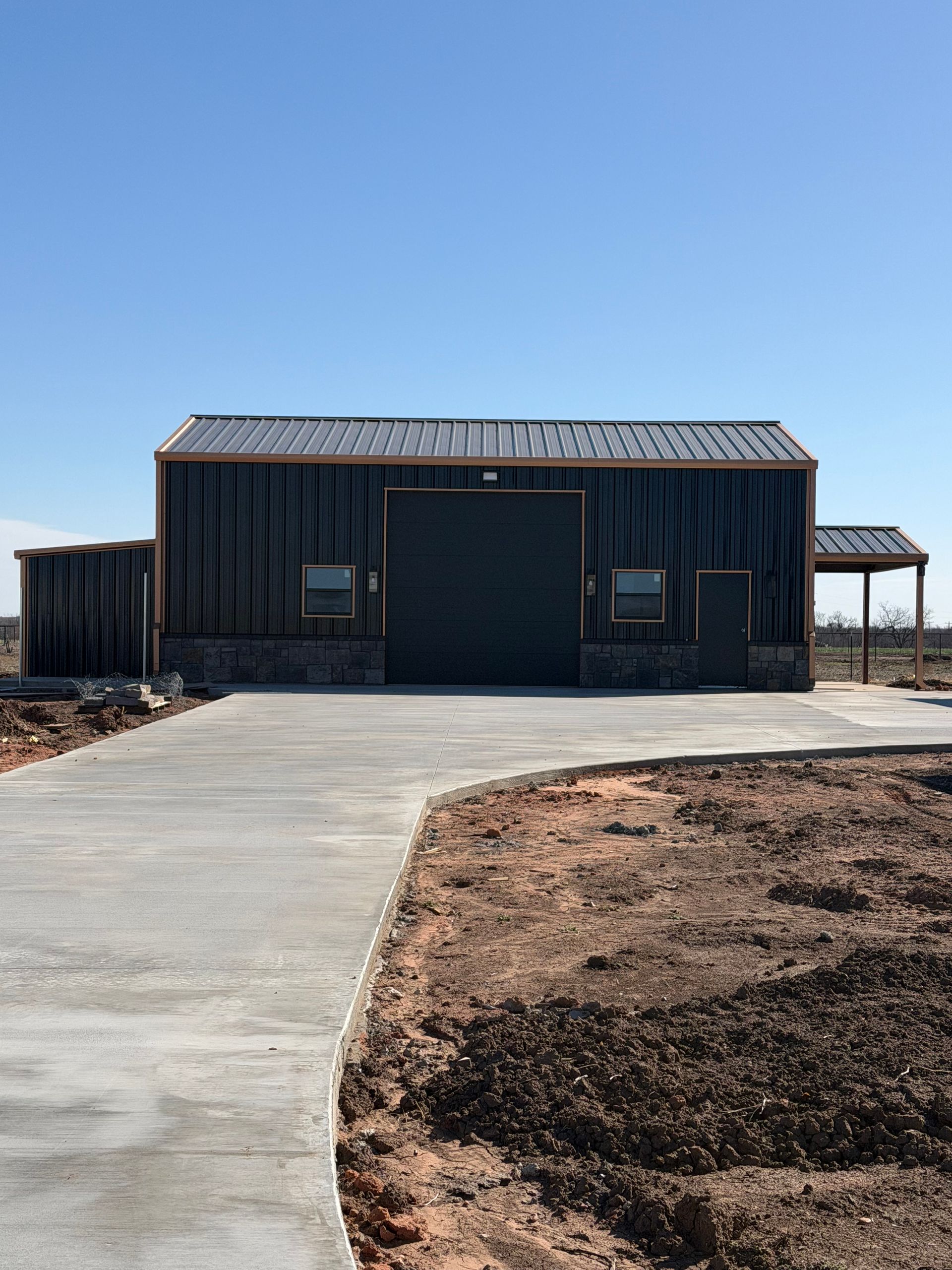 Dark modern barn-style building with a large garage door beside a curved concrete driveway under a clear blue sky