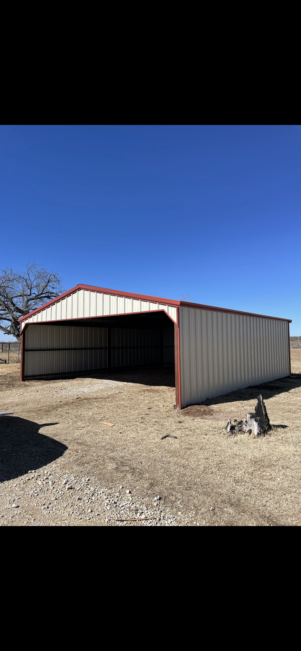 Open-sided metal shed with red roof on a gravel lot under a clear blue sky