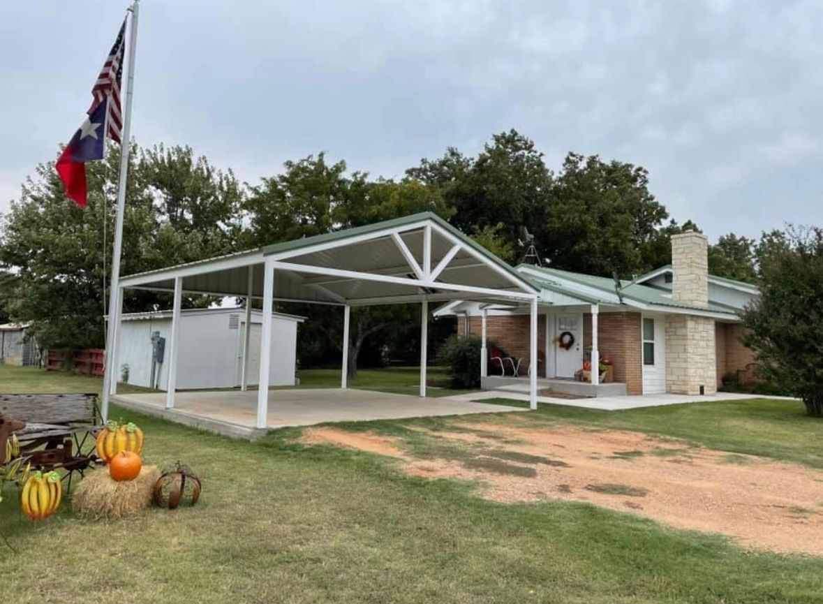 Rural house with white carport, Texas flag, and pumpkins on the lawn under cloudy skies
