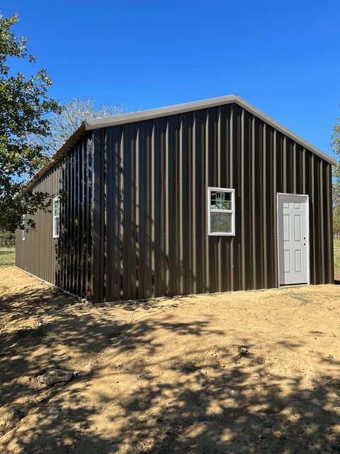 Dark brown metal shed with a white door and window, standing on a sunny dirt lot with trees nearby