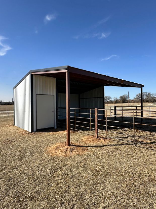Small white metal shed with a rusted covered pen in a dry field under a blue sky.