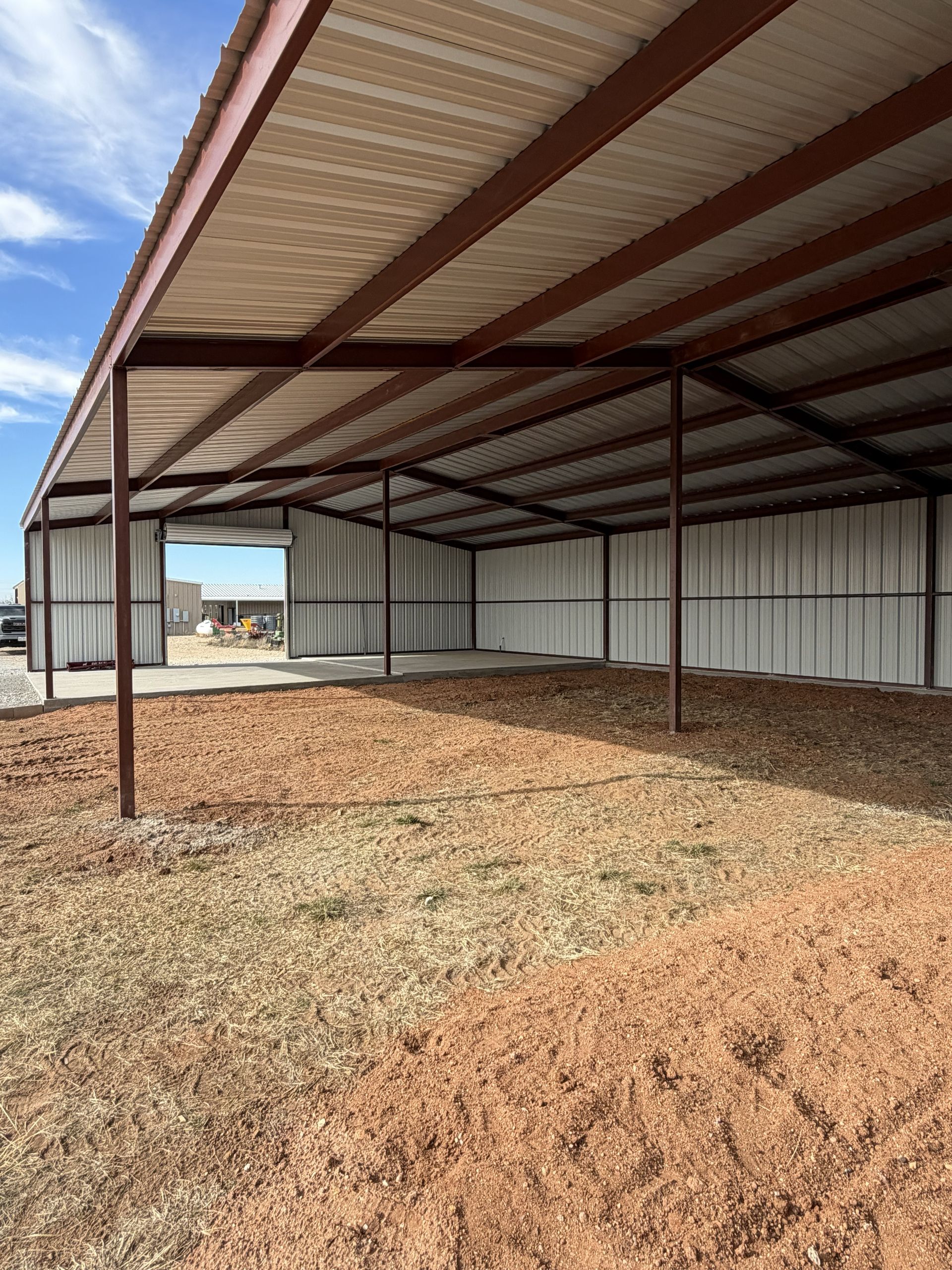 Open-sided metal shed with red beams and gravel ground under a cloudy sky