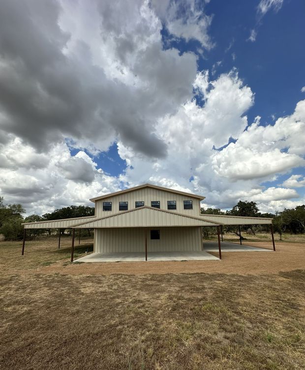 A small white elevated house in a dry field under a cloudy blue sky.