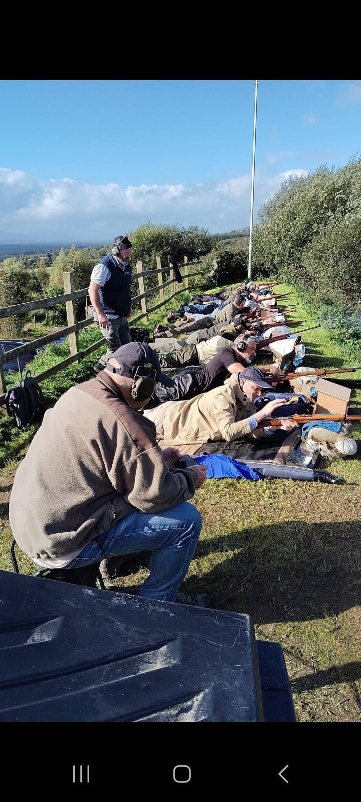 People at a shooting range, some lying down aiming rifles. Blue sky and foliage in the background.