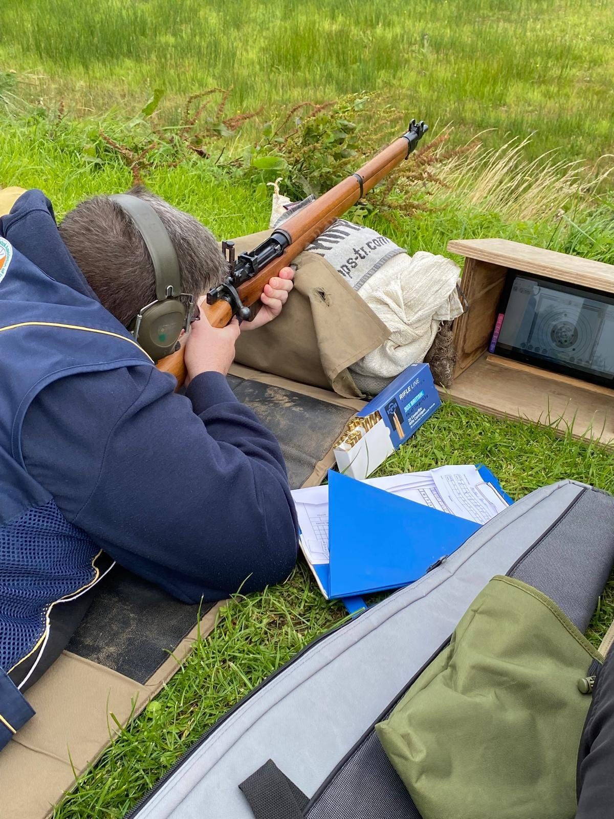 Person prone, aiming a rifle at a target on a grassy range. Gun case, ammunition, and target display visible.