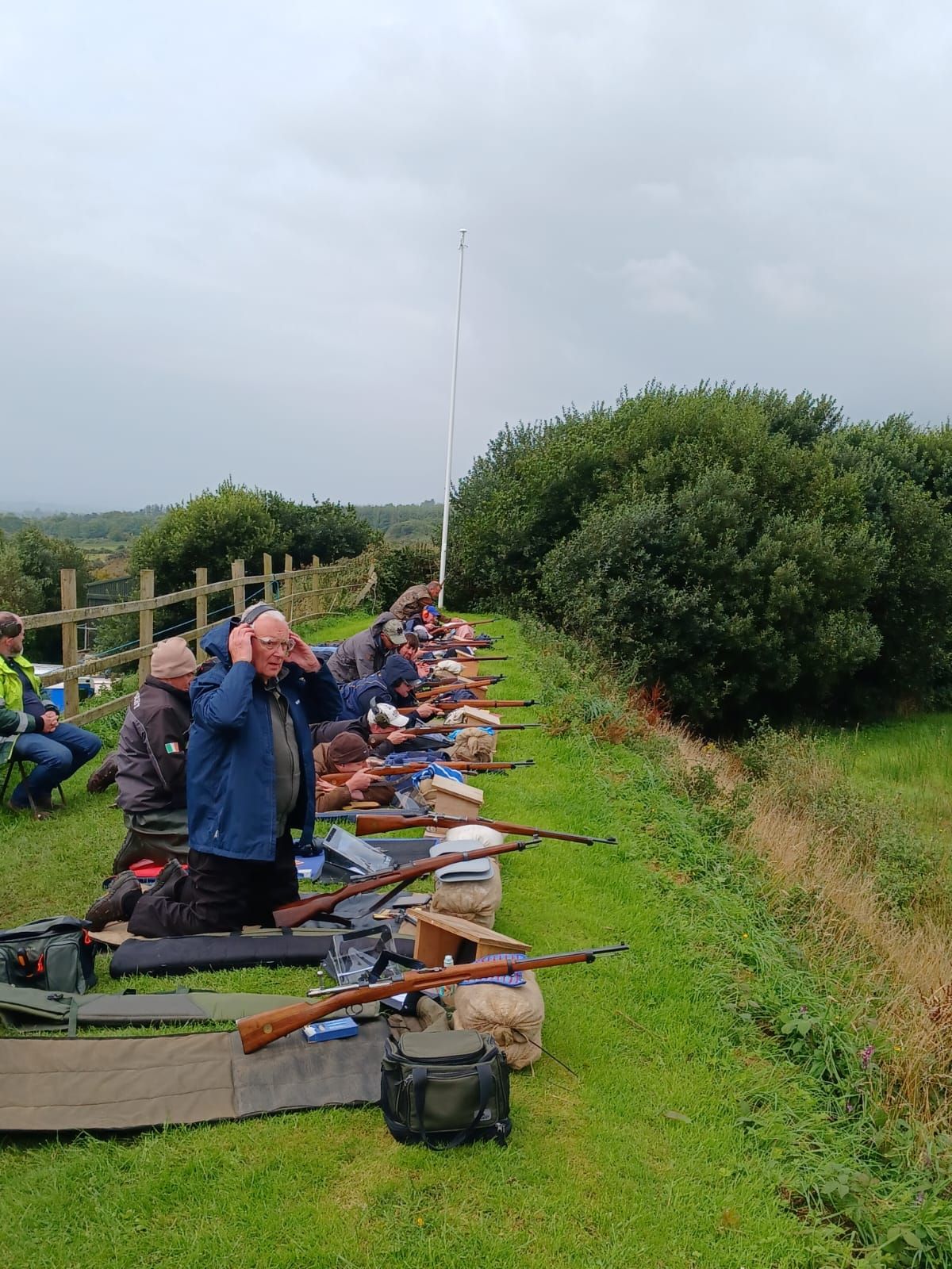 People at a shooting range, aiming rifles. Grassy embankment with cloudy sky. One man covers ears.