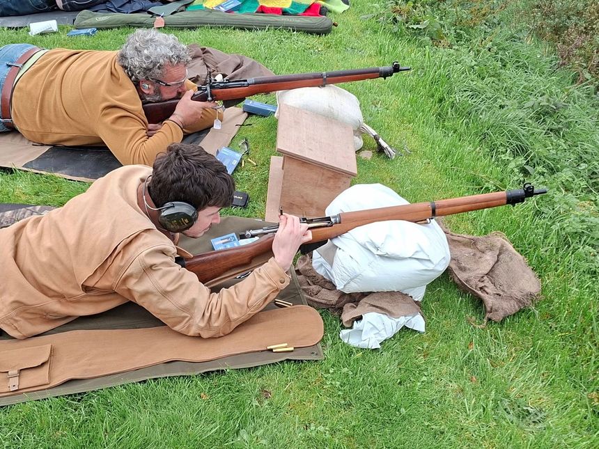 Two people aiming rifles on a grassy field. Both are lying down, focused, and using sandbags for support.