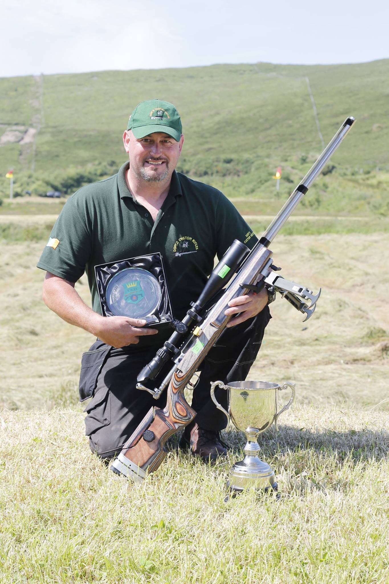 Man kneeling on grass, holding a rifle, a trophy, and a plaque. He wears a green shirt and a hat outdoors.