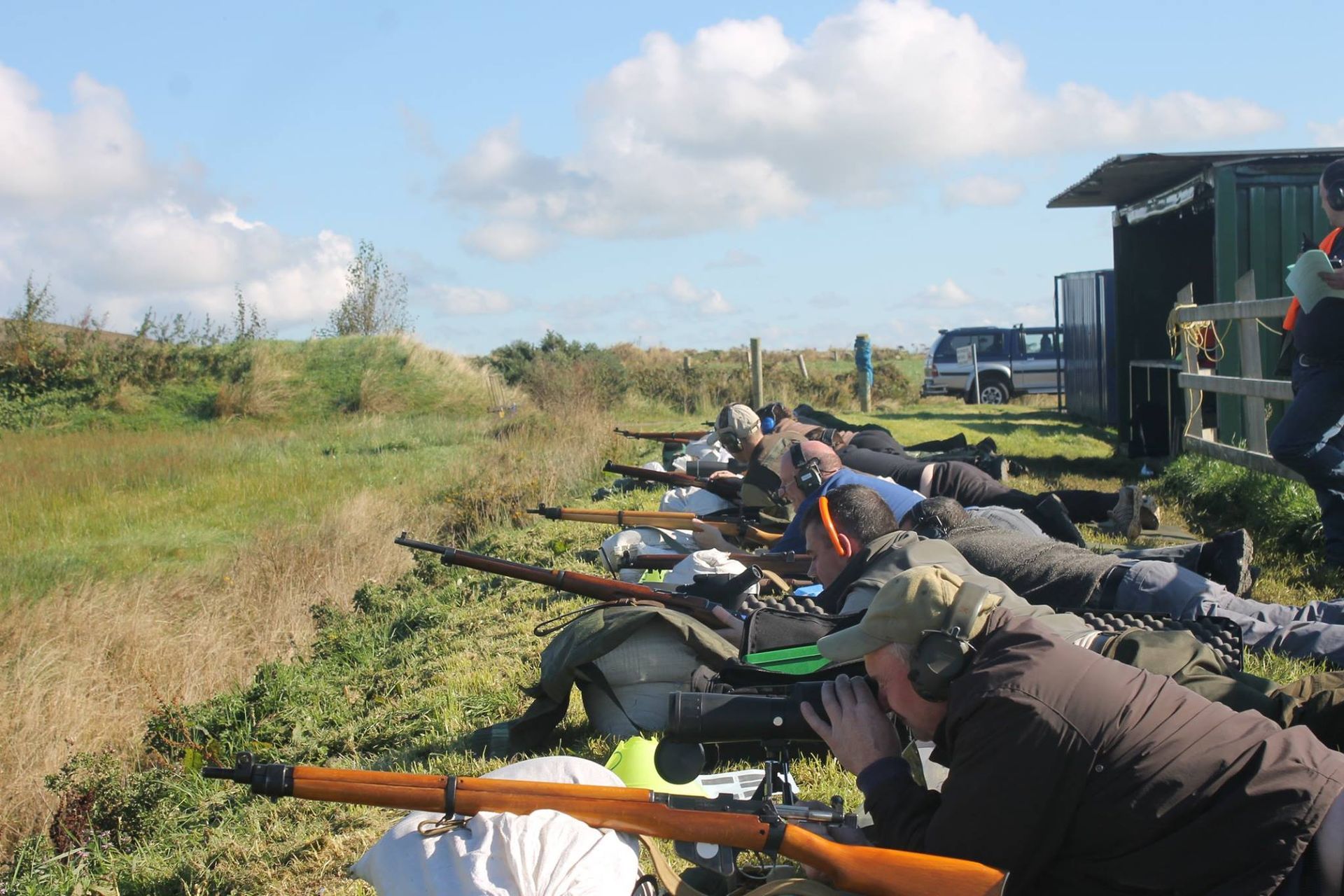People prone on the ground, aiming rifles at a target outdoors on a grassy field. A blue sky with clouds in the background.