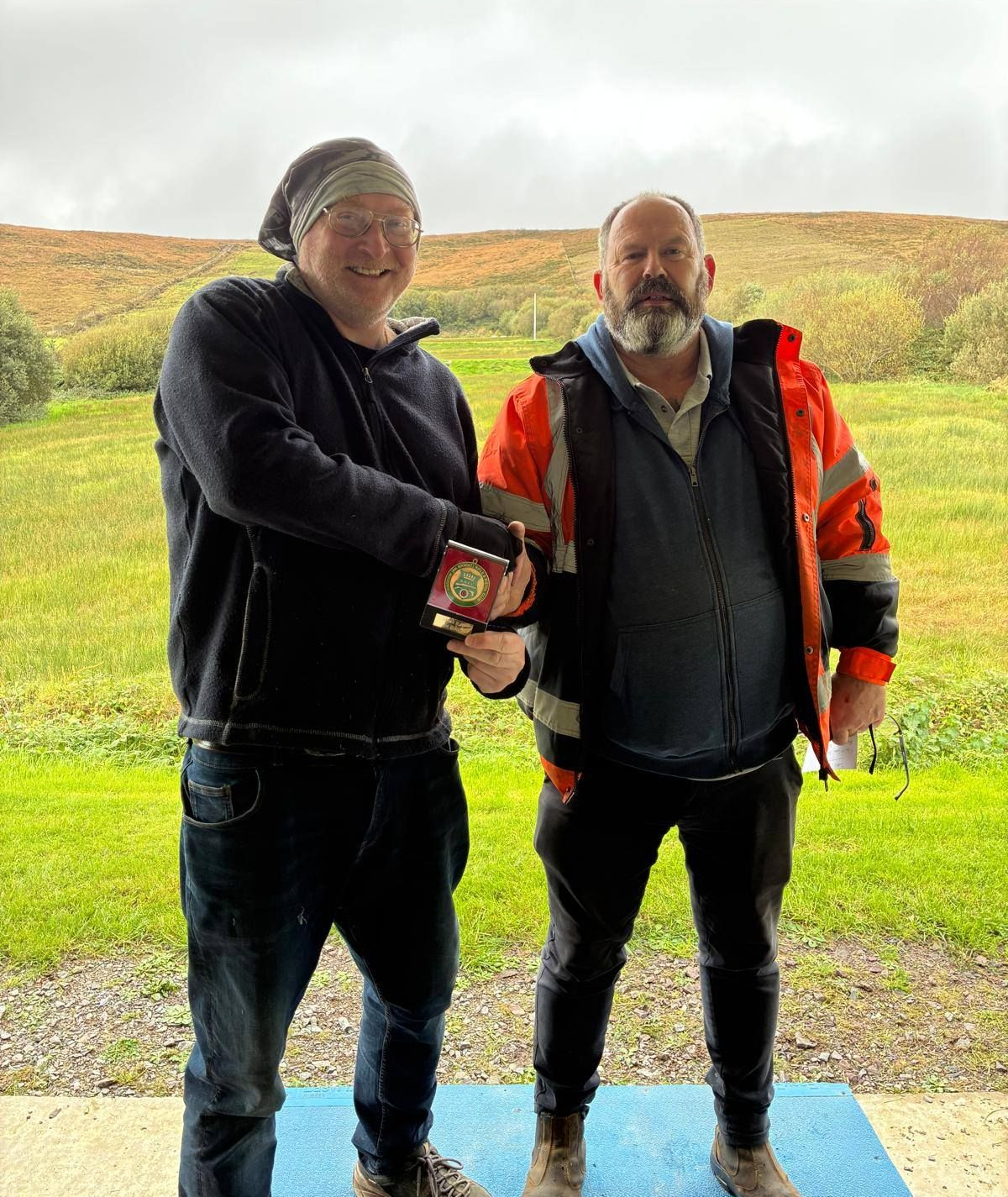 Two men outdoors, one handing a small trophy to the other. Man on left wearing a head covering.