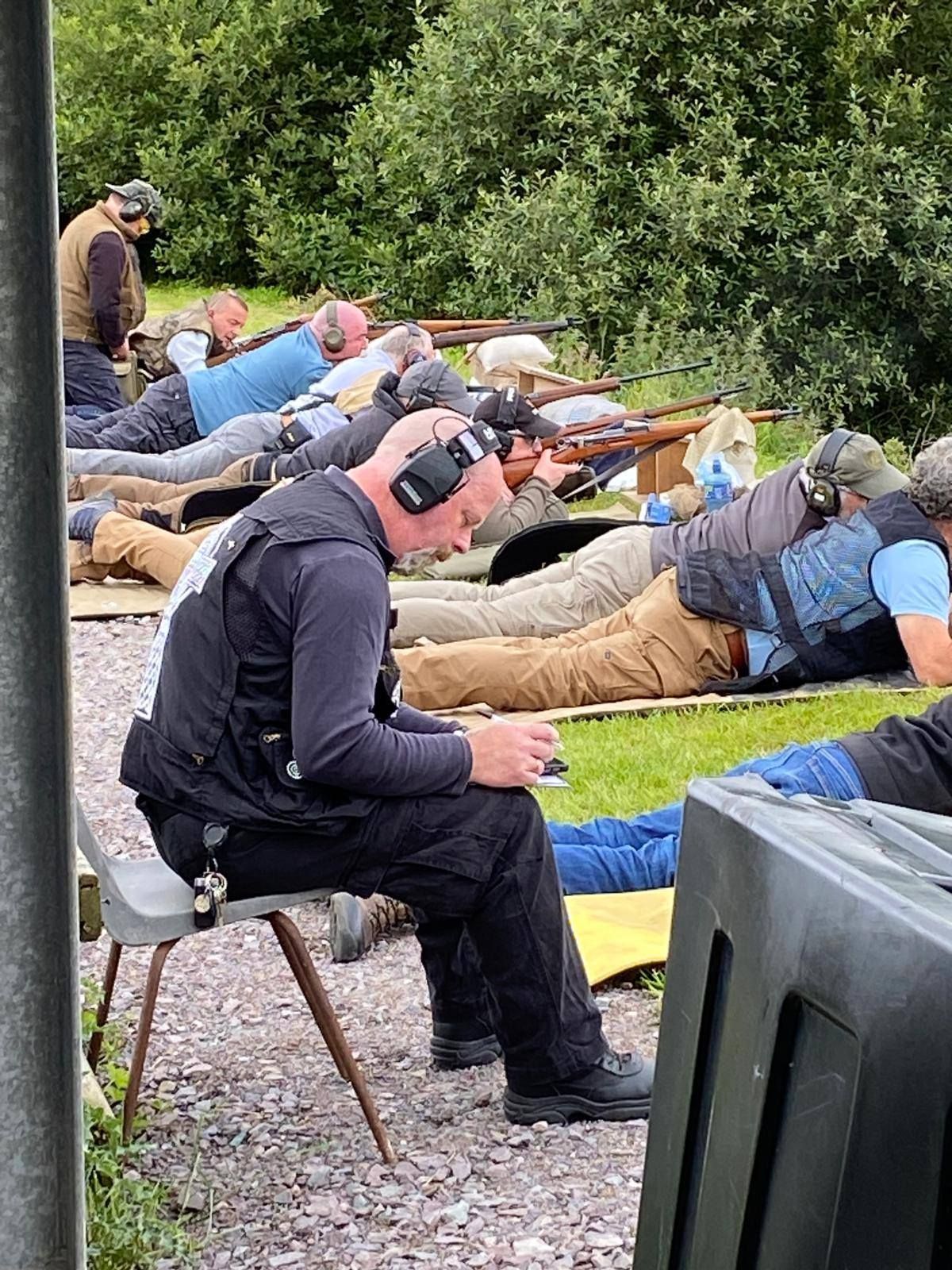 People in shooting range, some prone, one seated. Person in black vest looks down at device. Green grass, trees.