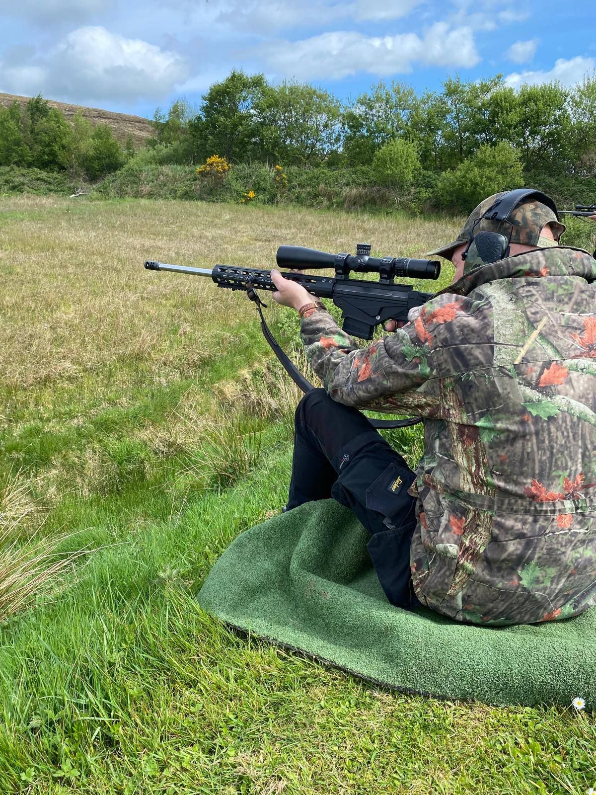 Person in camouflage jacket aiming a rifle at a shooting range, seated on a green mat.