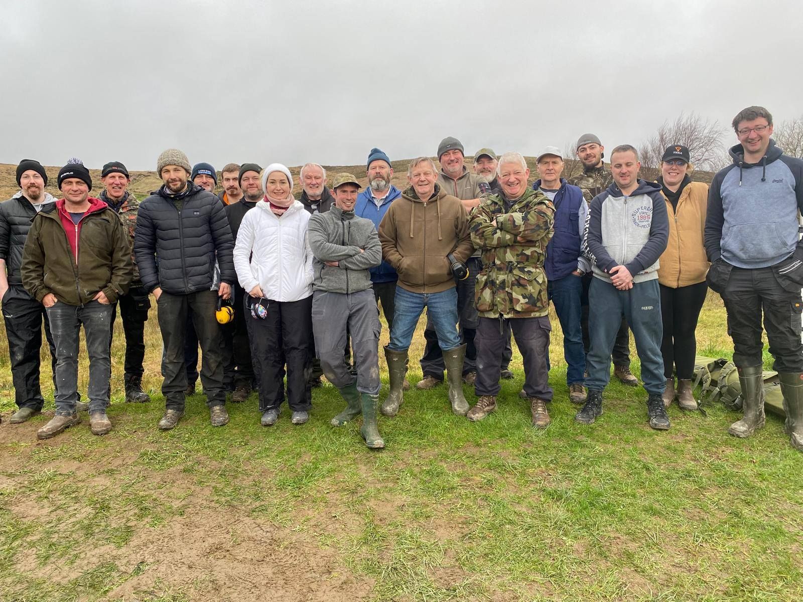 Group of men standing on grassy field on overcast day. Most wear casual clothes, some have hats, and boots.