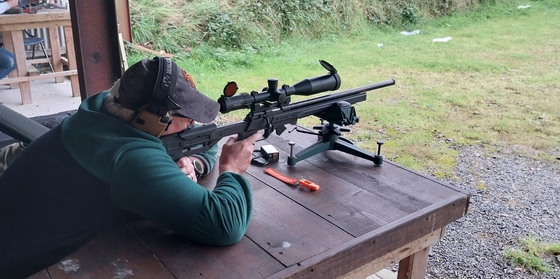 Person aims a rifle with a scope, resting on a table at an outdoor shooting range. Green grass in the background.