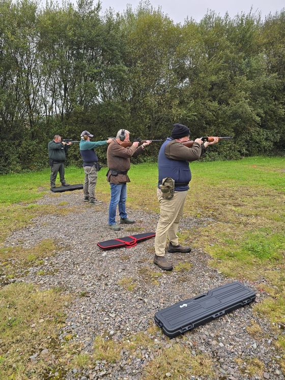 Four people aiming rifles at a shooting range, outdoors. Trees in the background. Gun cases on the ground.