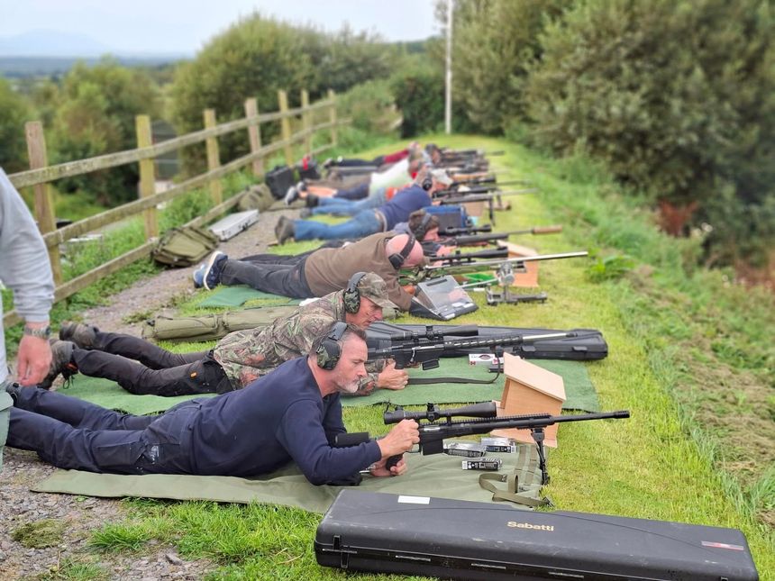 People prone, aiming rifles at a shooting range. Green grass, wooden fence, overcast sky.