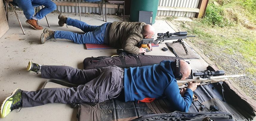 Two people are prone, aiming rifles. A third person sits nearby. They are outdoors near a grassy area.