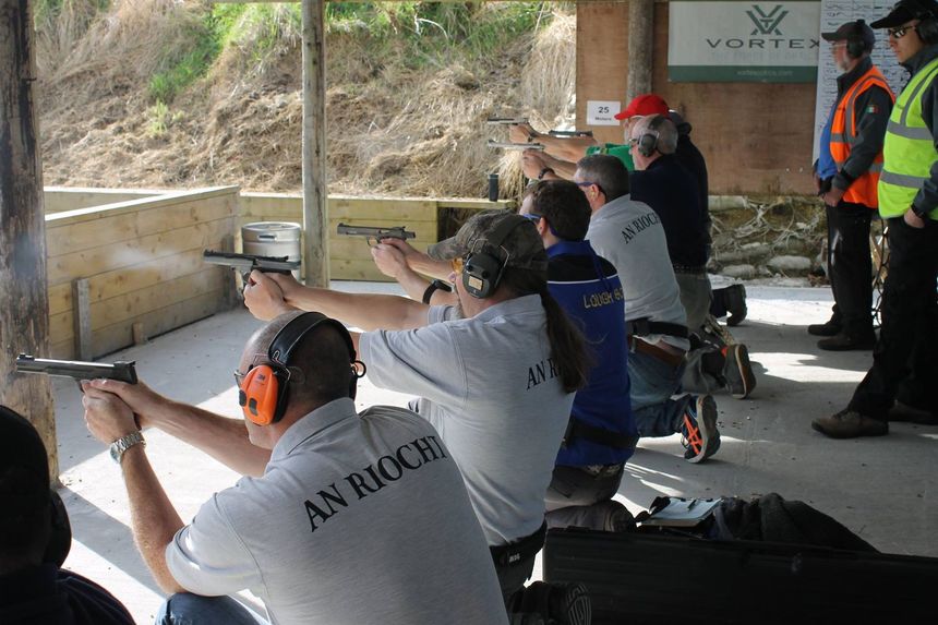 People shooting handguns at an outdoor range. Shooters are kneeling, wearing ear protection, and aiming at targets.