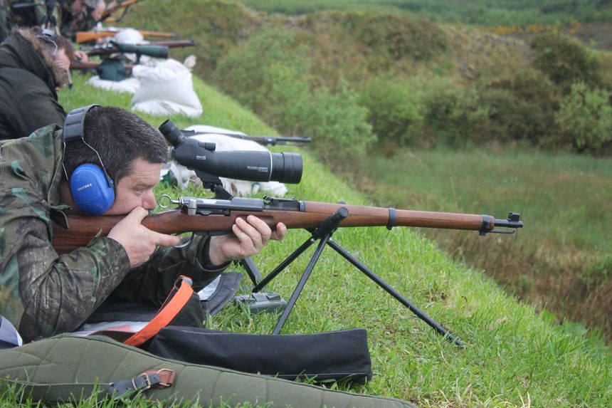 Man aiming rifle at a shooting range, wearing ear protection. Rifle is on a stand, green grass, and other shooters .