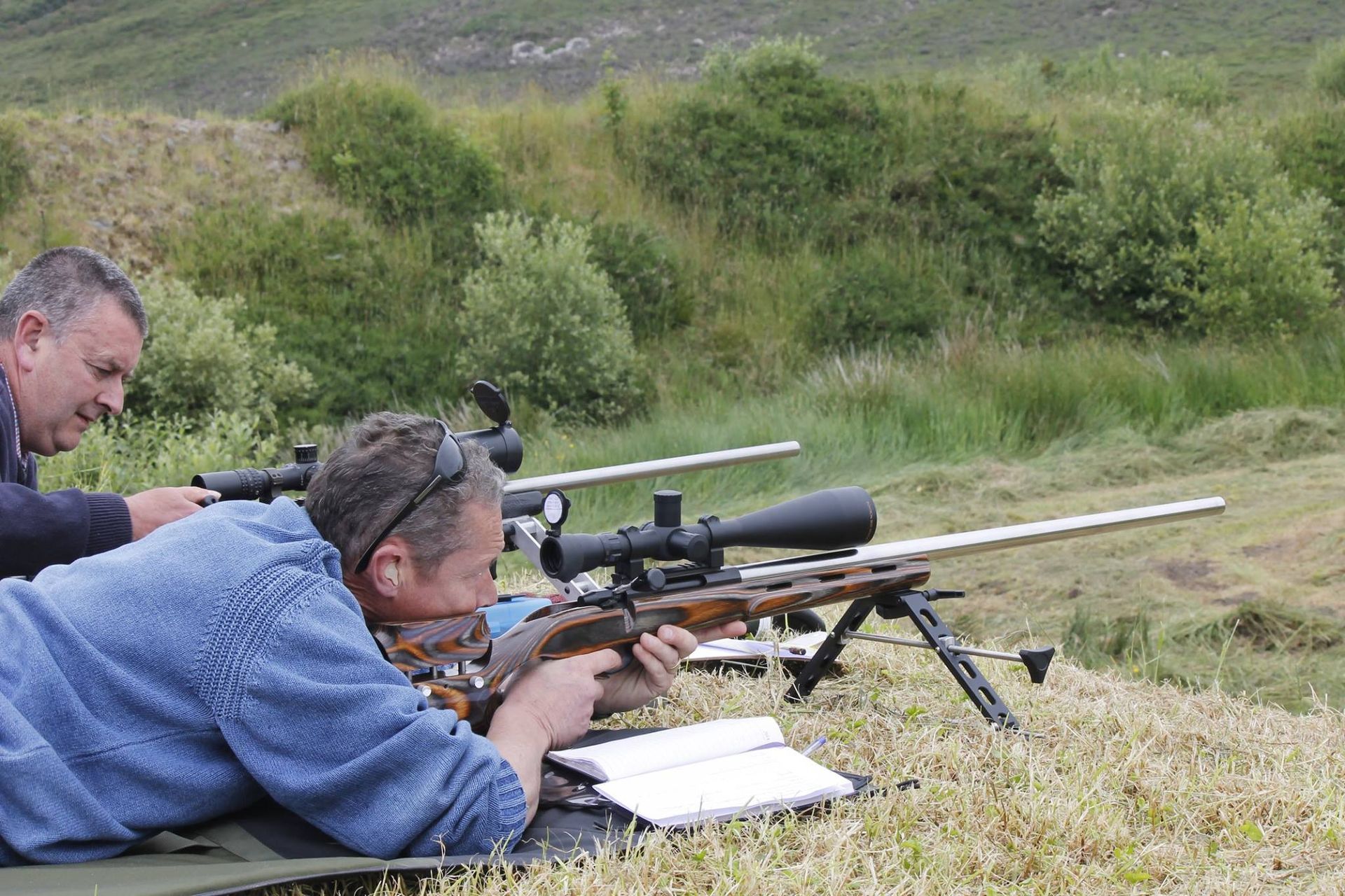 Two men aiming rifles at a shooting range. Man in blue shirt, focused, prone position. Outdoors, grassy hillside.