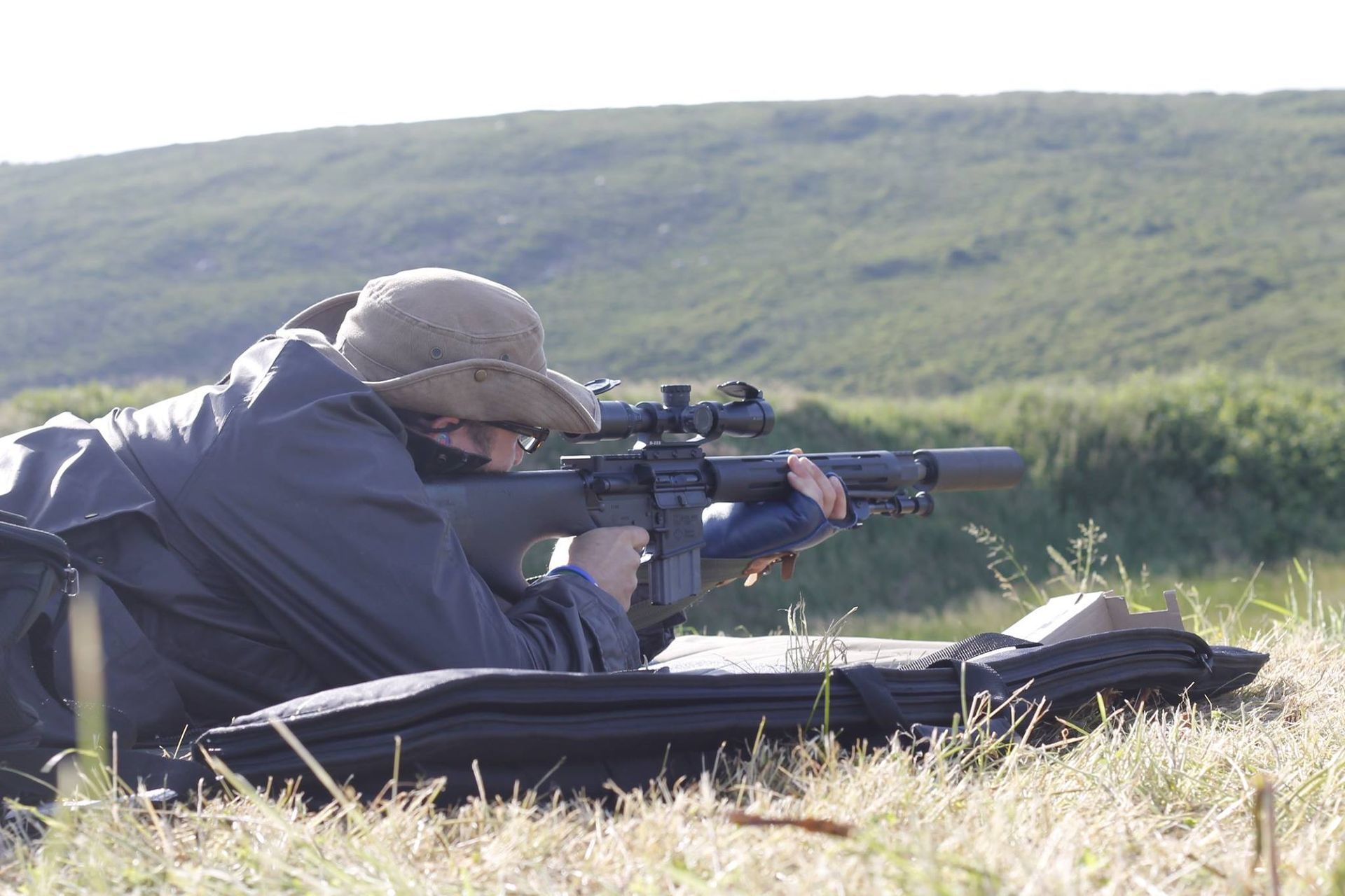 Person aiming a rifle with a scope, lying prone in grassy field, wearing a hat and jacket.