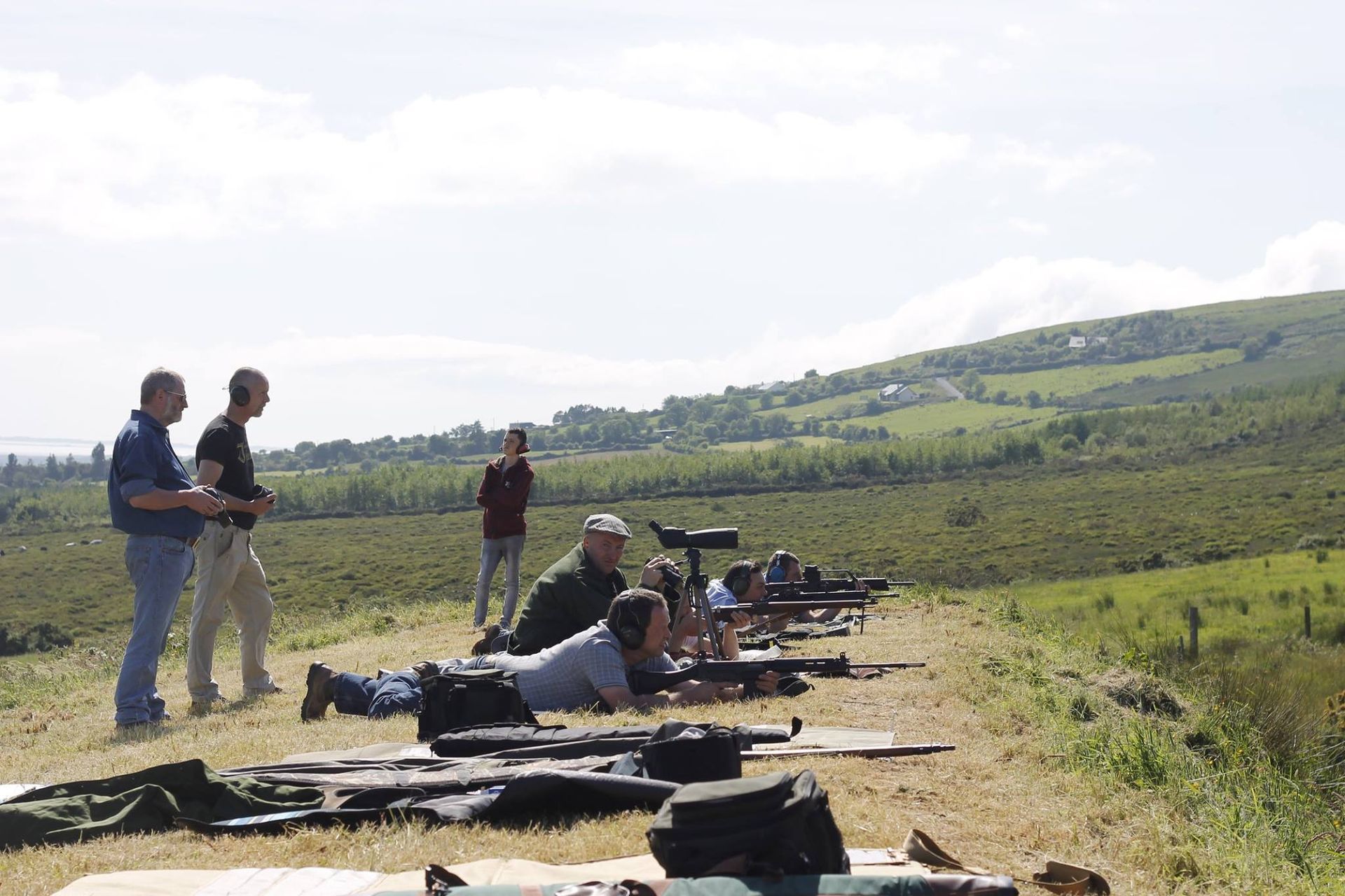 People shooting rifles at a firing range with a grassy landscape in the background.