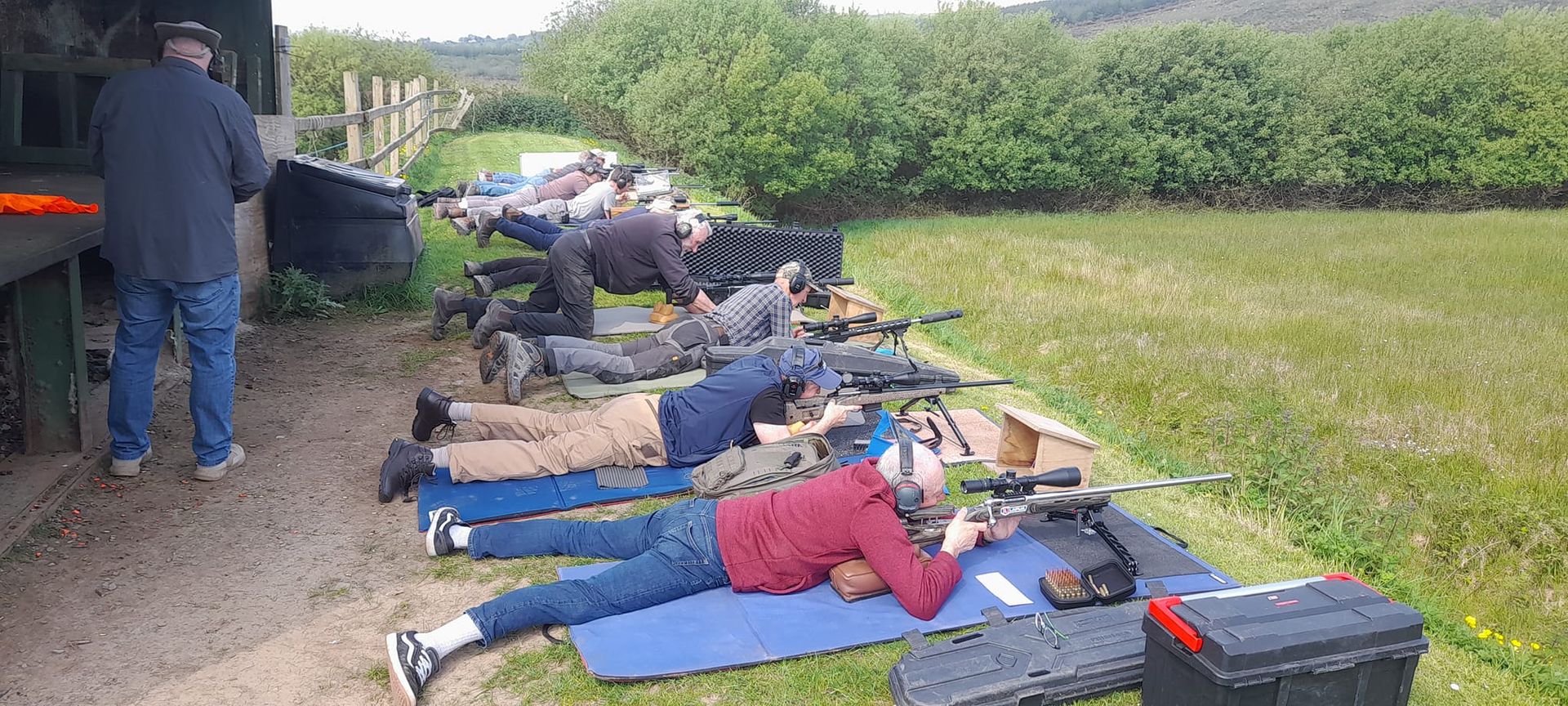People lying prone, aiming rifles at a shooting range. A man stands nearby.
