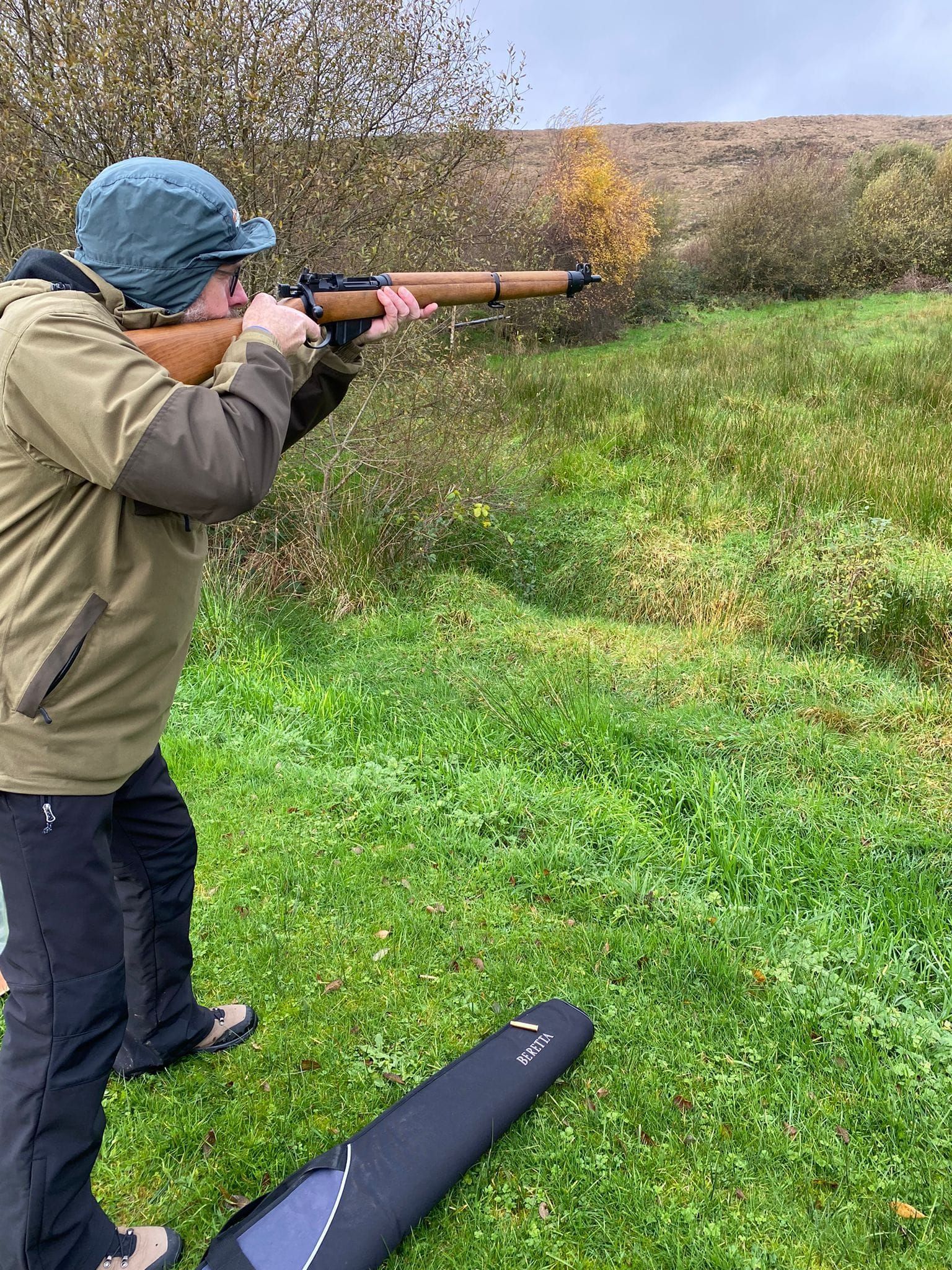Person aiming a rifle on a grassy hillside, wearing outdoor clothing and a hat, with a rifle case in the foreground.