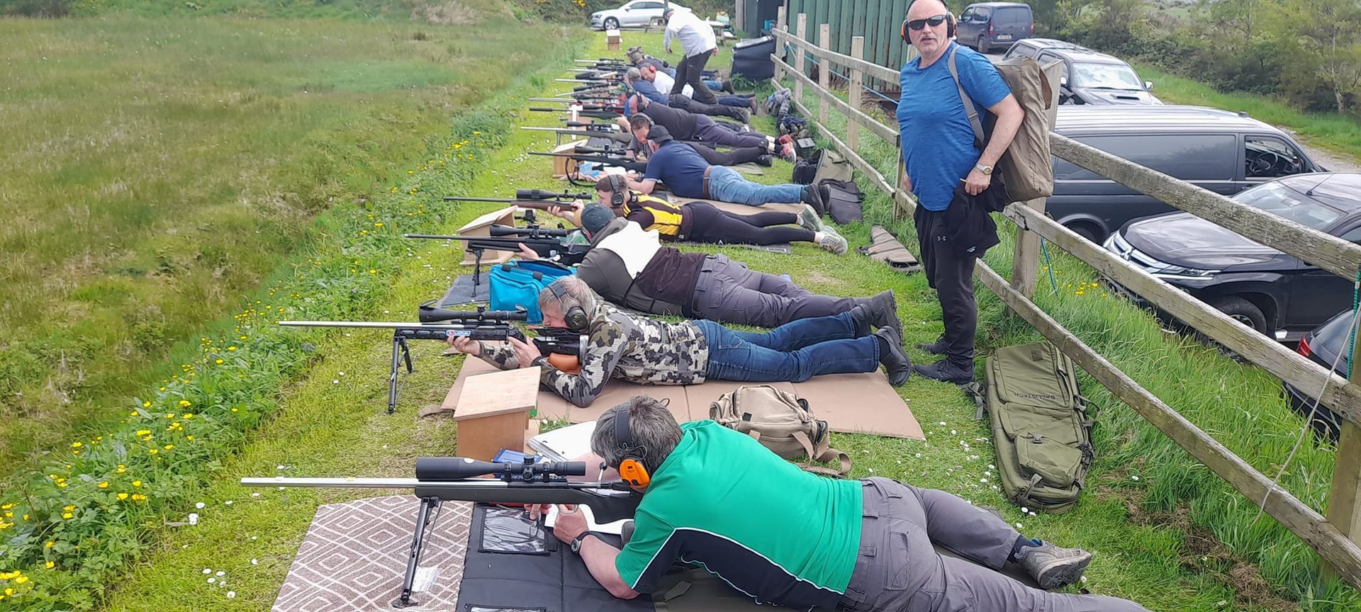 People prone on a firing range, aiming rifles. A man stands beside them. Grass and vehicles are nearby.