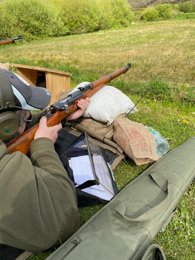 Person aiming a rifle outdoors, using a support rest. Green grass and a wooden structure are visible.