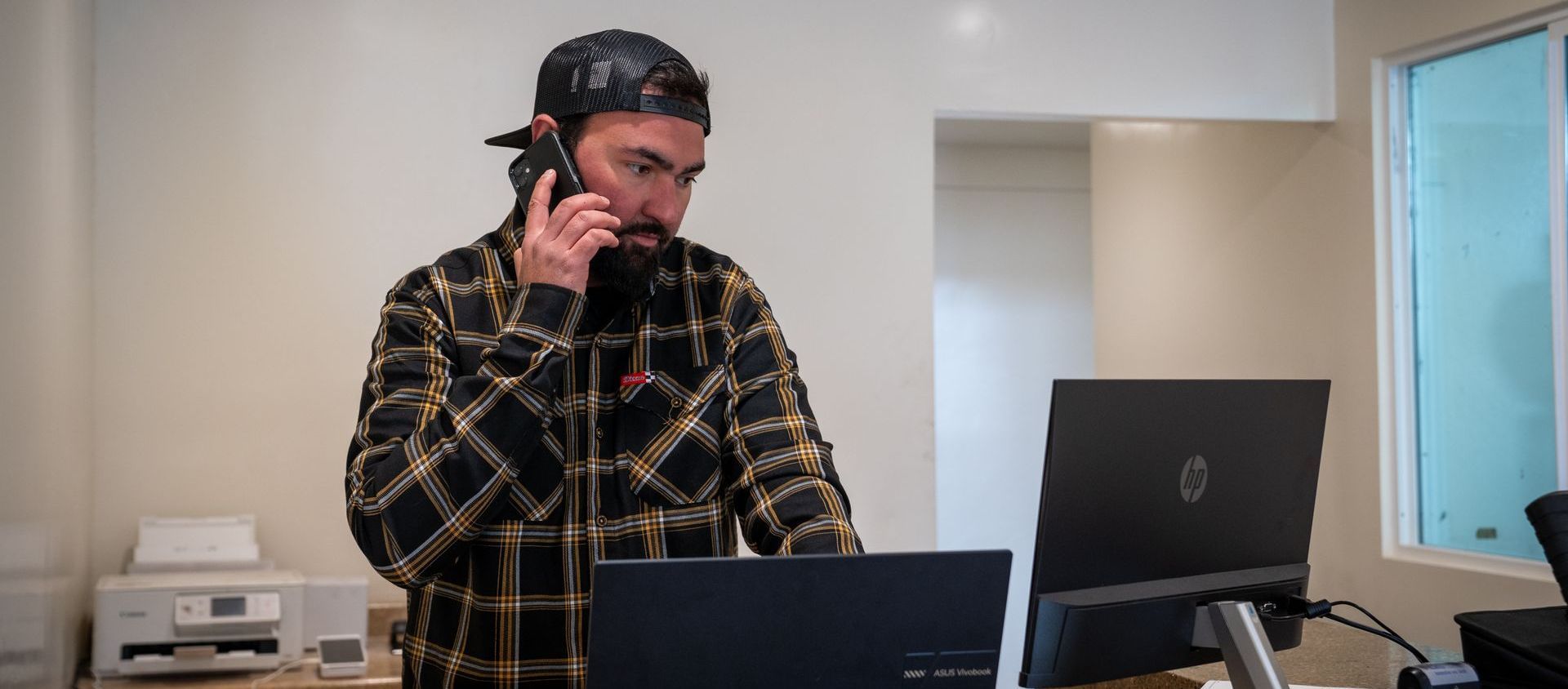 Man in a plaid shirt talking on a phone beside a laptop and monitor in a bright office