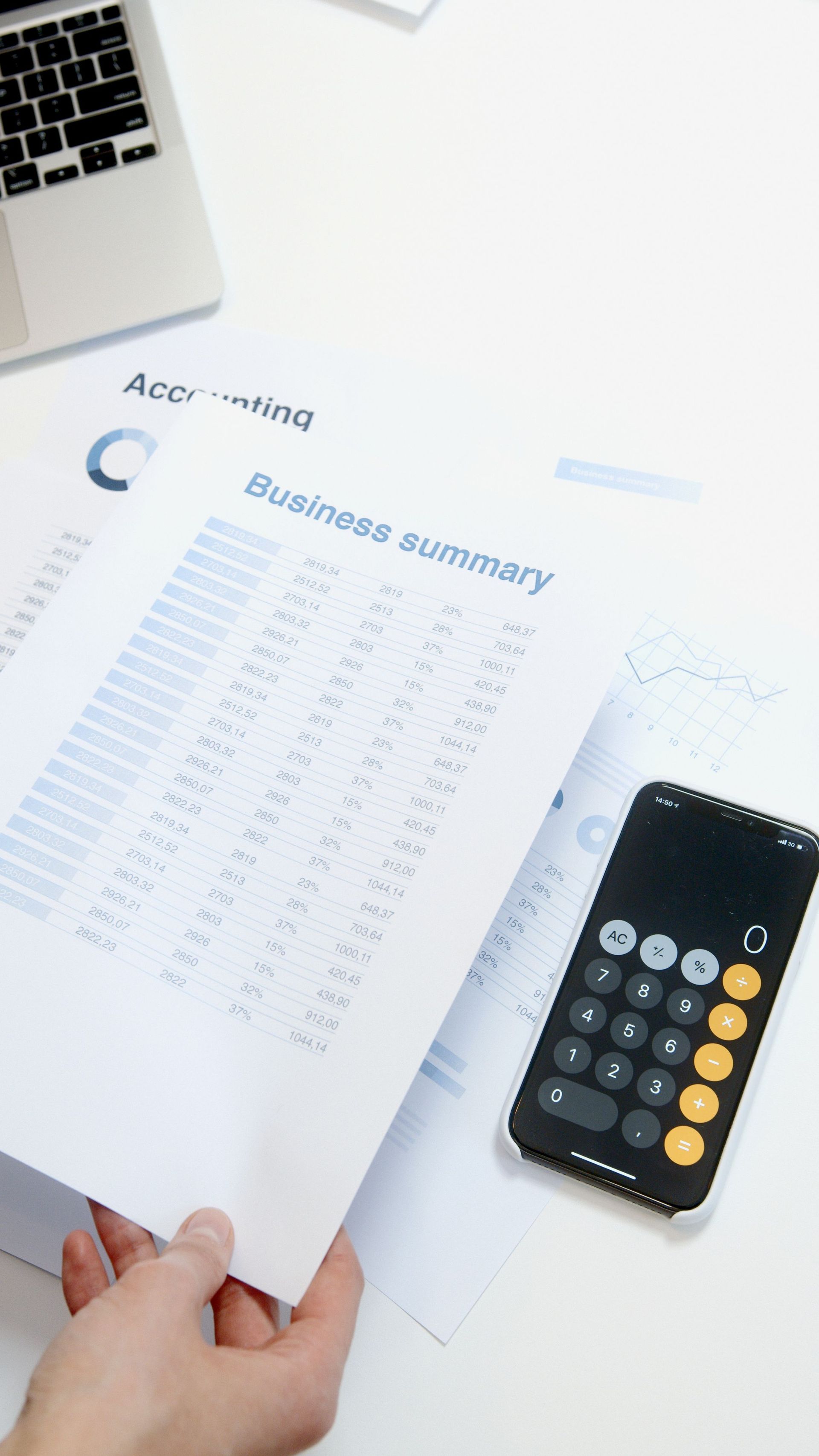 Hand holding a budget summary sheet beside a calculator on a white desk with a laptop corner visible