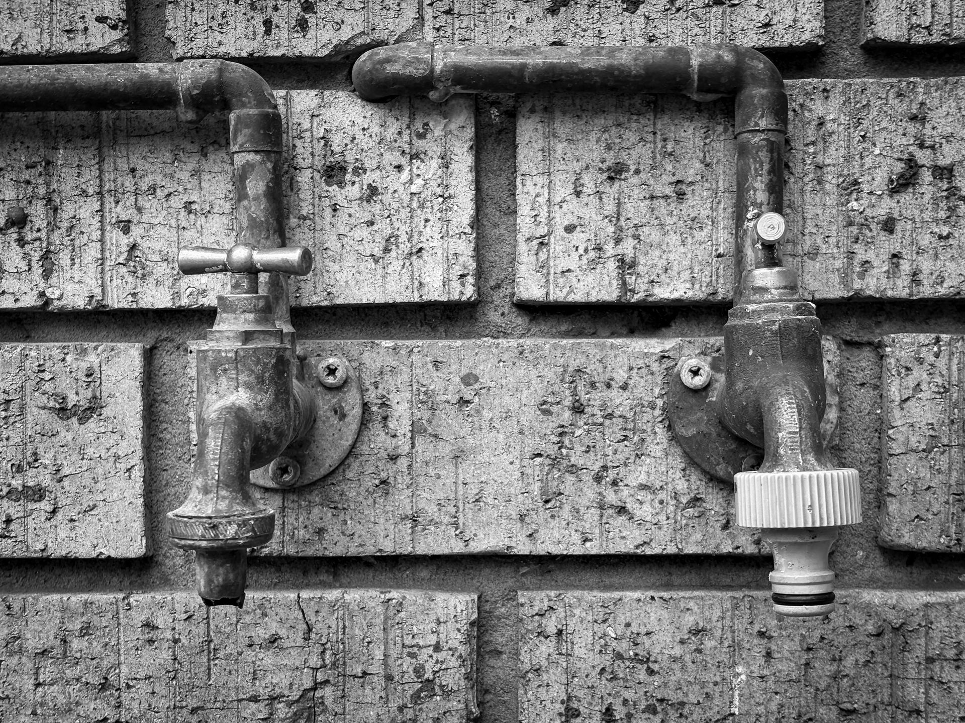 Two weathered outdoor water faucets on a brick wall in black and white.