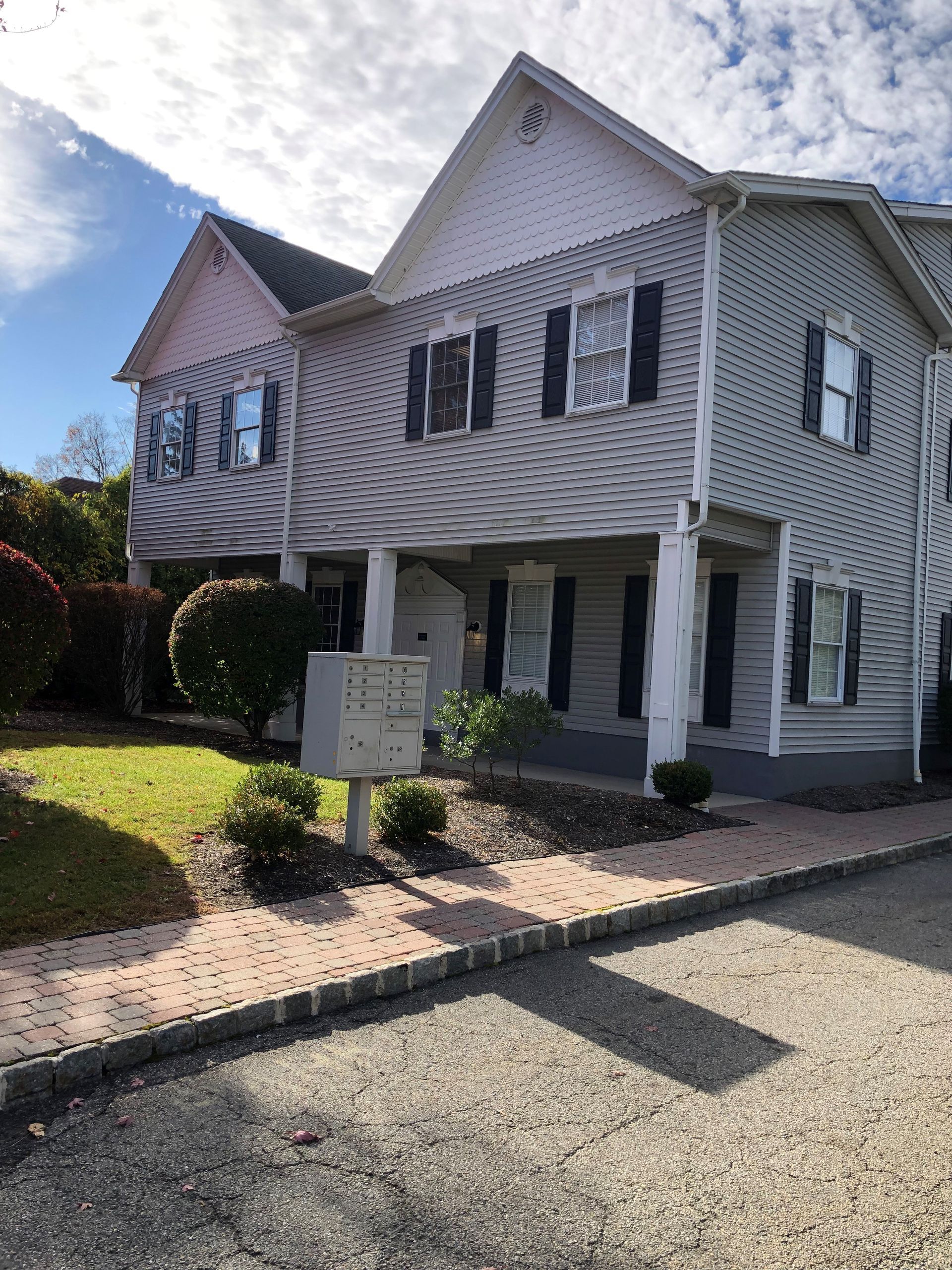 Two-story light gray house with black shutters, mailbox in front, sidewalk, green grass, and blue sky.