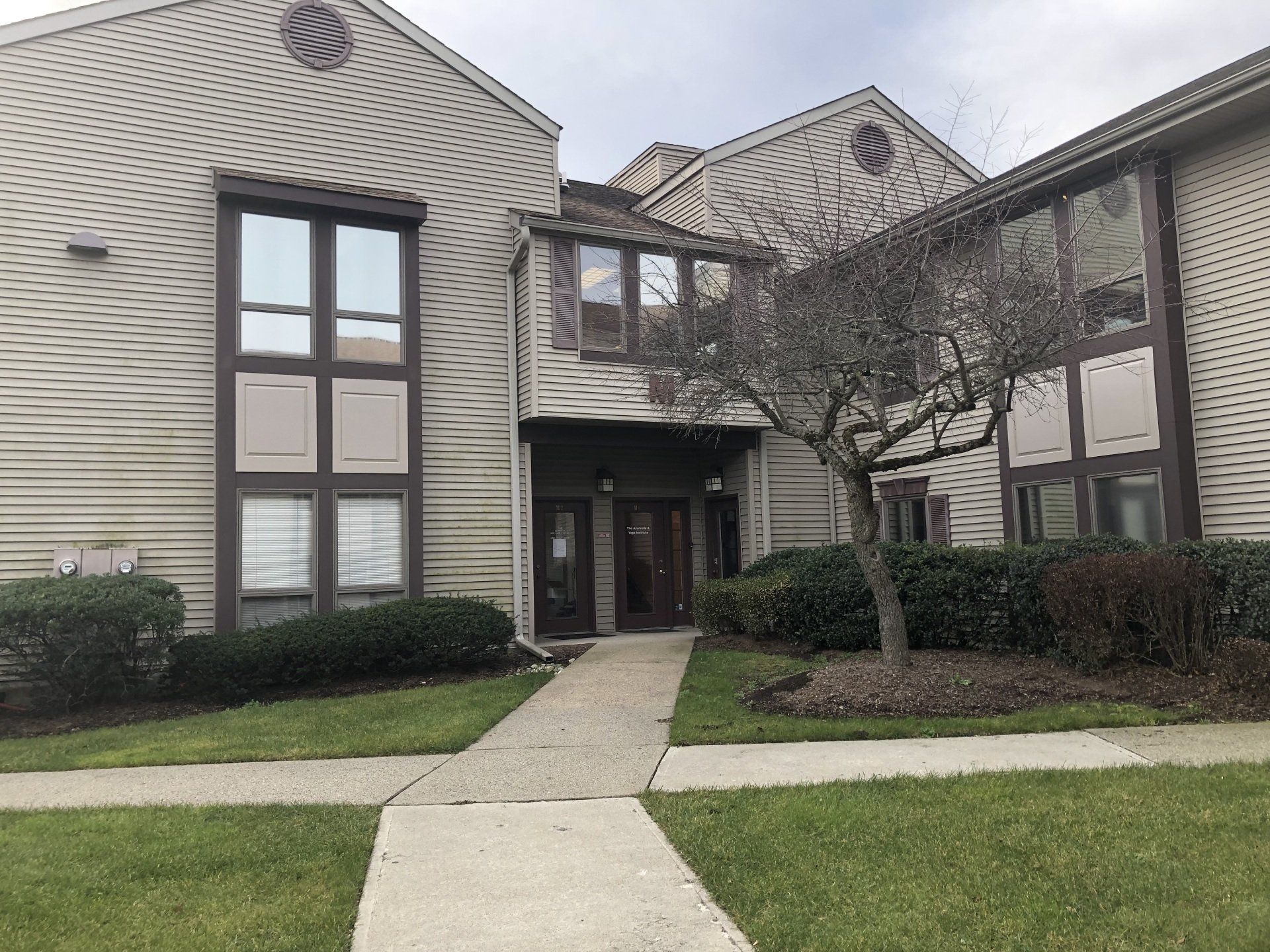 Beige multi-unit building with brown trim, windows, and a bare tree in front. Concrete path leads to the front doors.