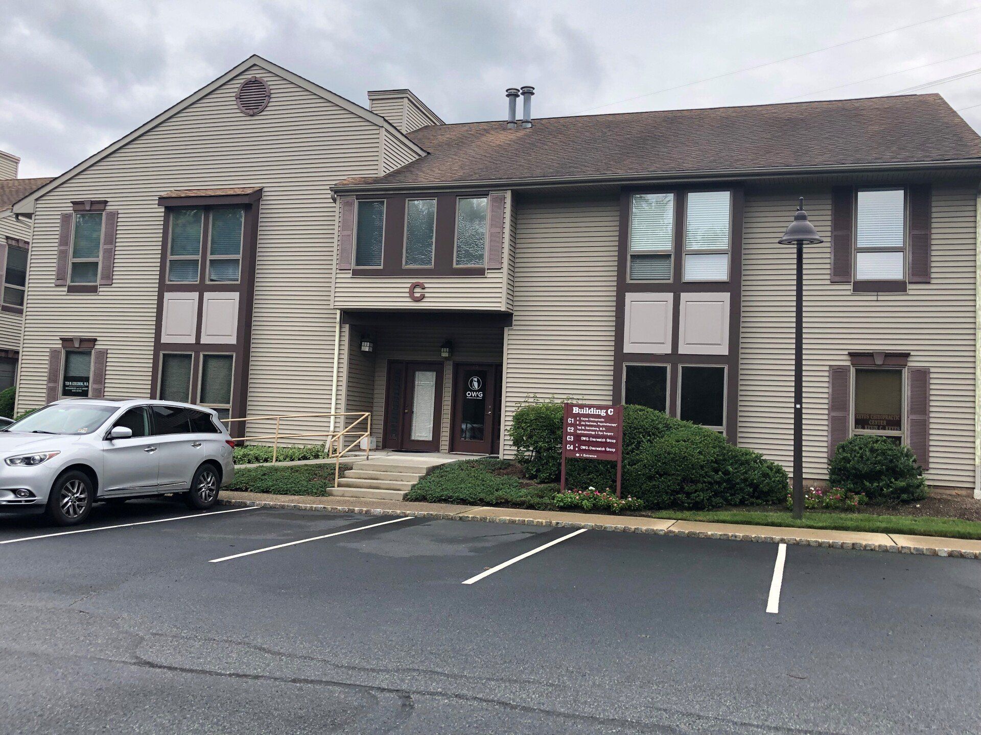 Two-story building with tan siding, brown trim, and a small parking lot. A silver SUV is parked on the left.