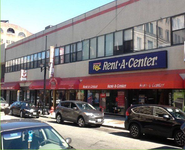 Rent-A-Center storefront in a city setting, with cars parked out front under a red awning.