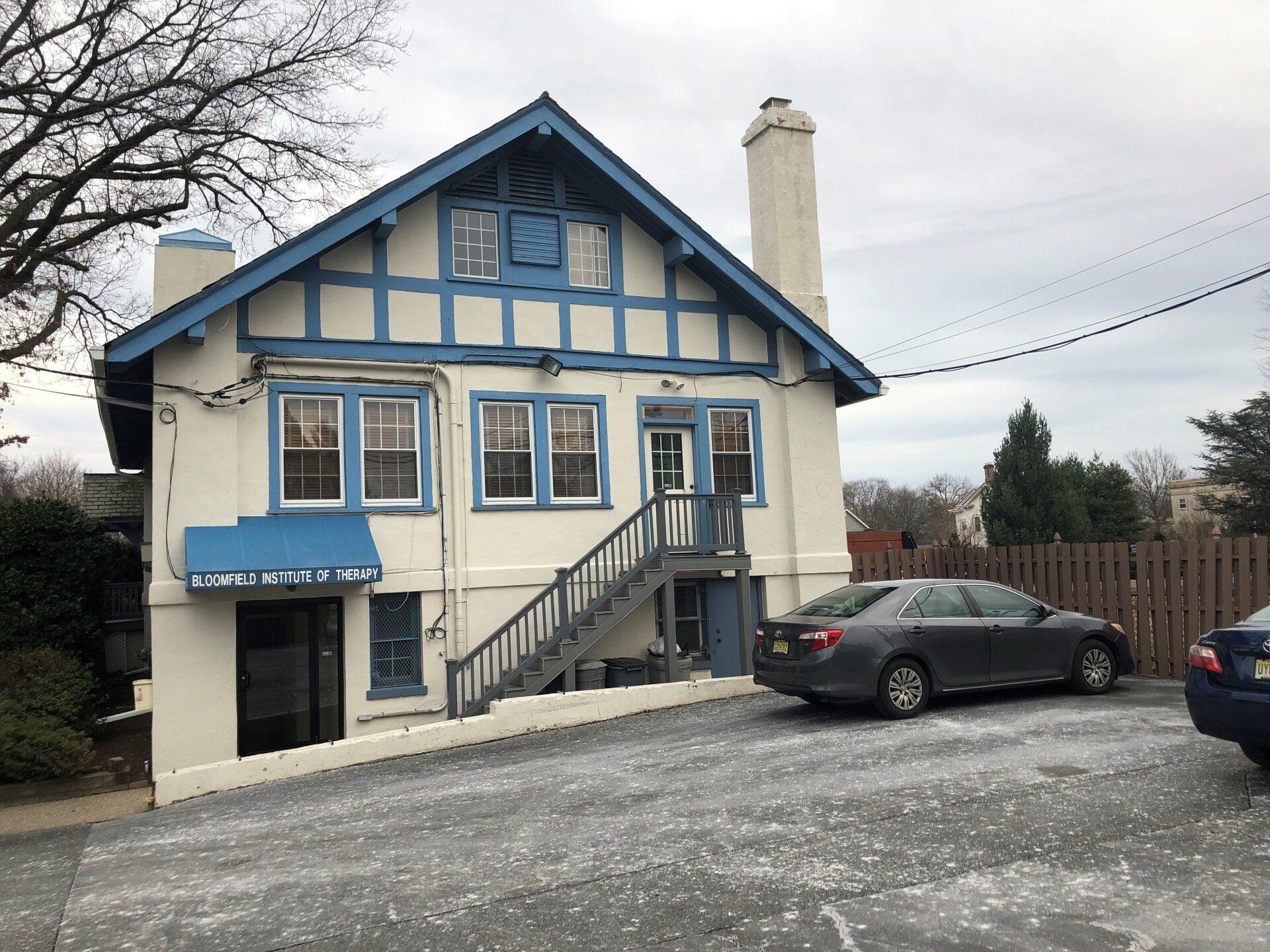 White house with blue trim, staircase, and parked cars on a cloudy day.