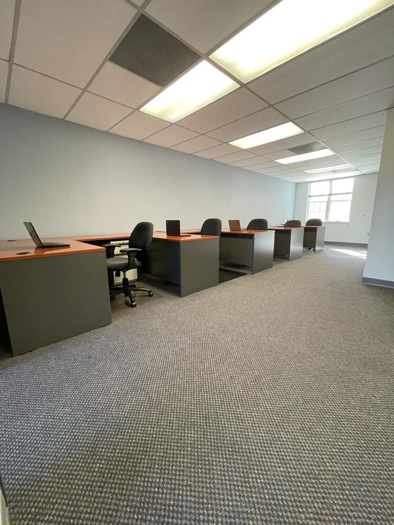 Empty office with desks, chairs, and carpet. Blue wall, ceiling lights, and a window in the background.