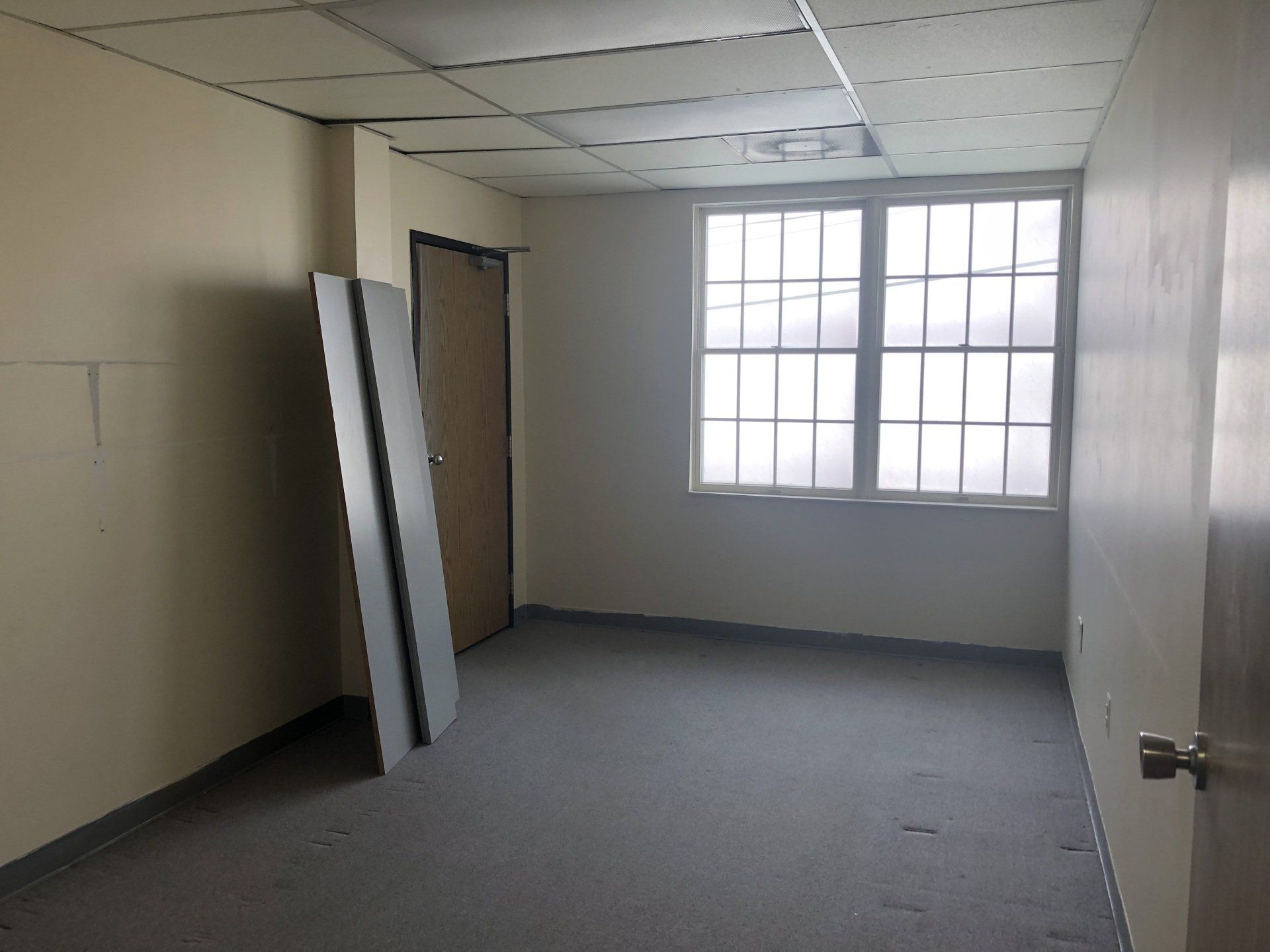 Empty room with a window, door, and gray carpet. Beige walls and white ceiling tiles.