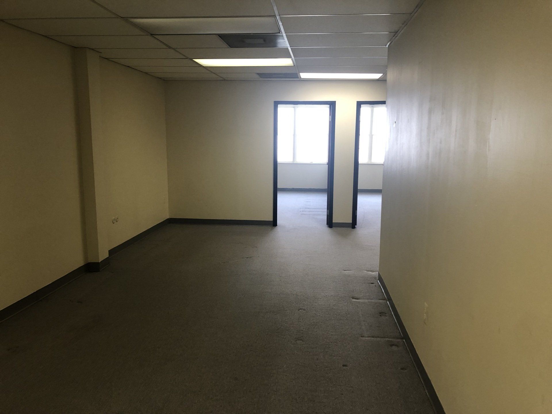 An empty, beige office suite with gray carpet, recessed ceiling lights, and two doorways leading to another room.