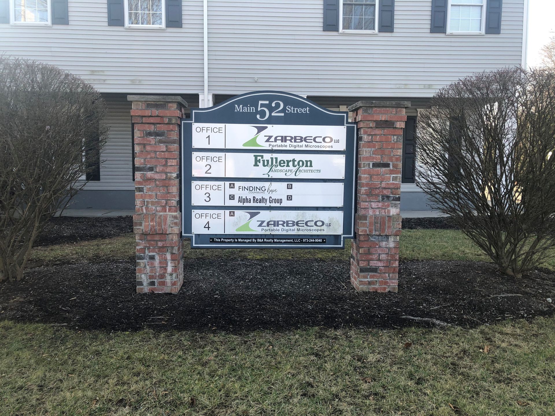 Sign with business names at 52 Elm Street, framed by brick columns, surrounded by landscaping.