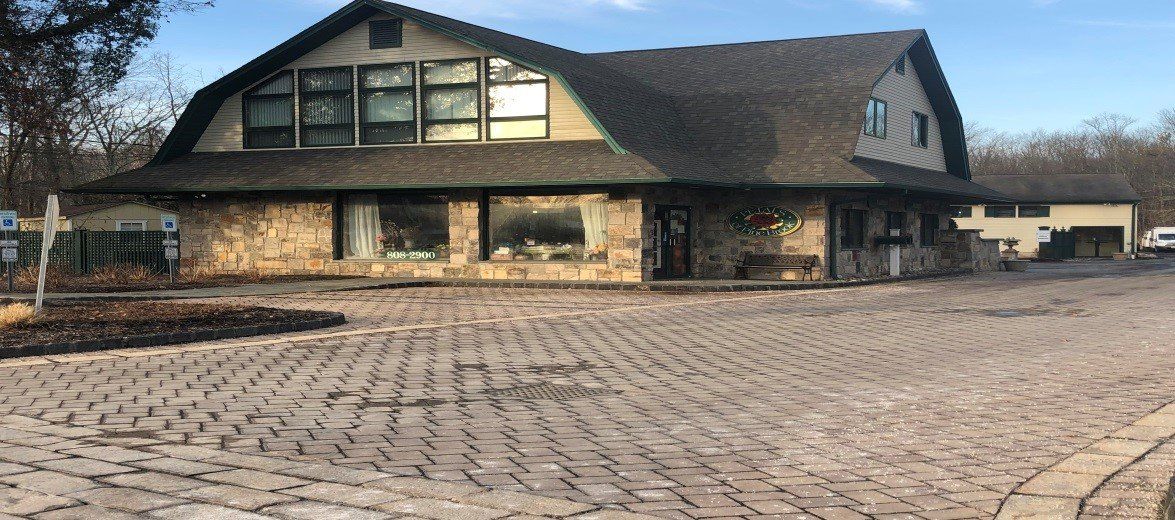 A stone building with a large gambit roof and dark shingled siding, situated behind a wide, patterned brick driveway.
