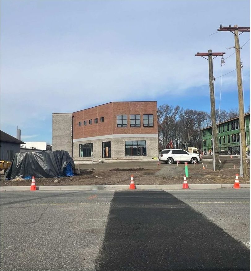 New building under construction with brick and gray stone facade, orange cones in the foreground, white SUV nearby.