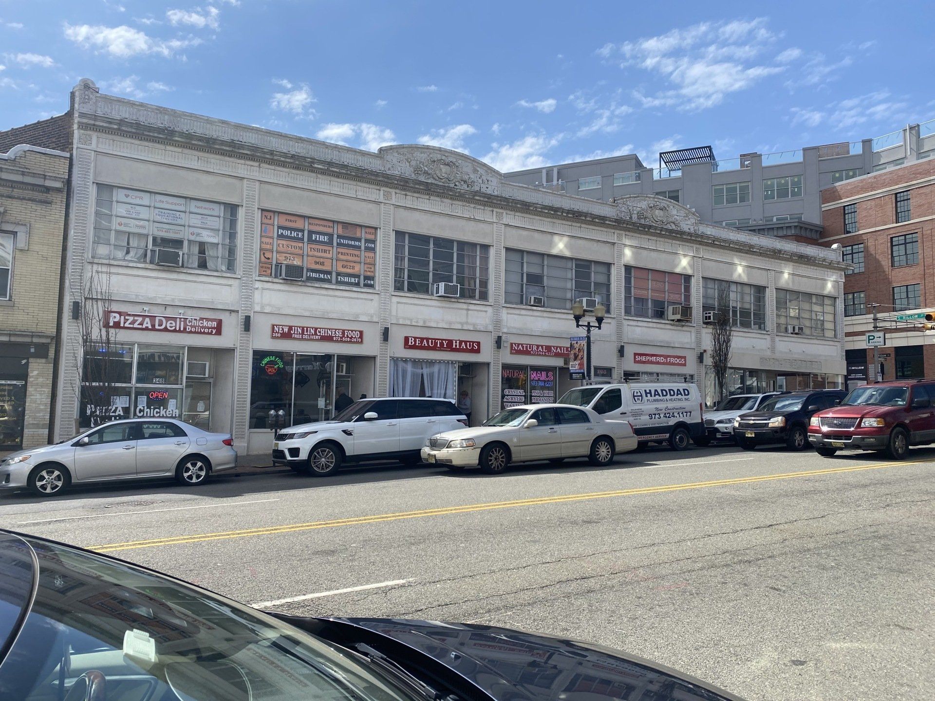 Two-story white building with storefronts. Cars parked in front on a city street under a blue sky.