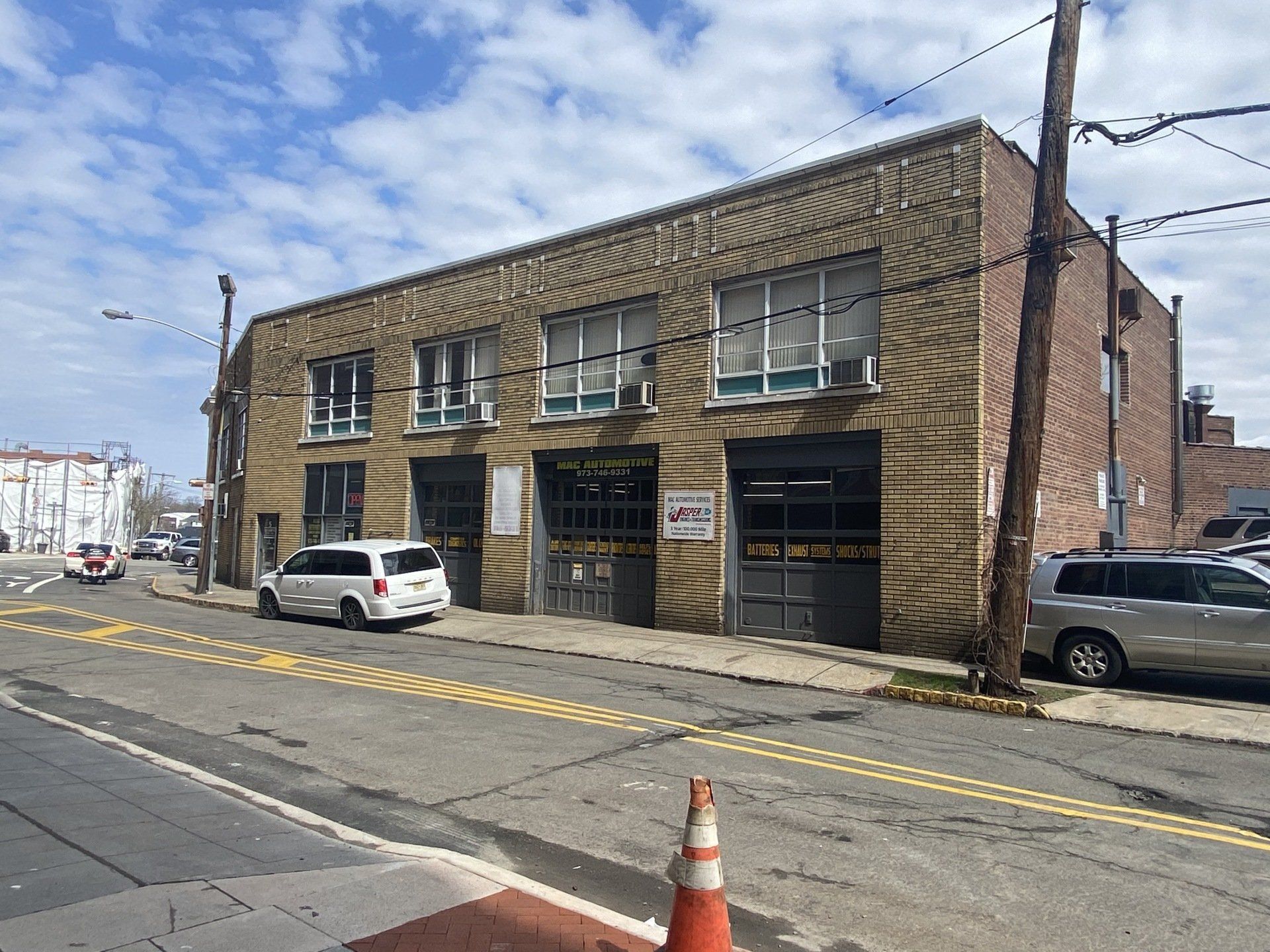 Two-story brick building with garage doors, parked cars, and street in front under a partly cloudy sky.