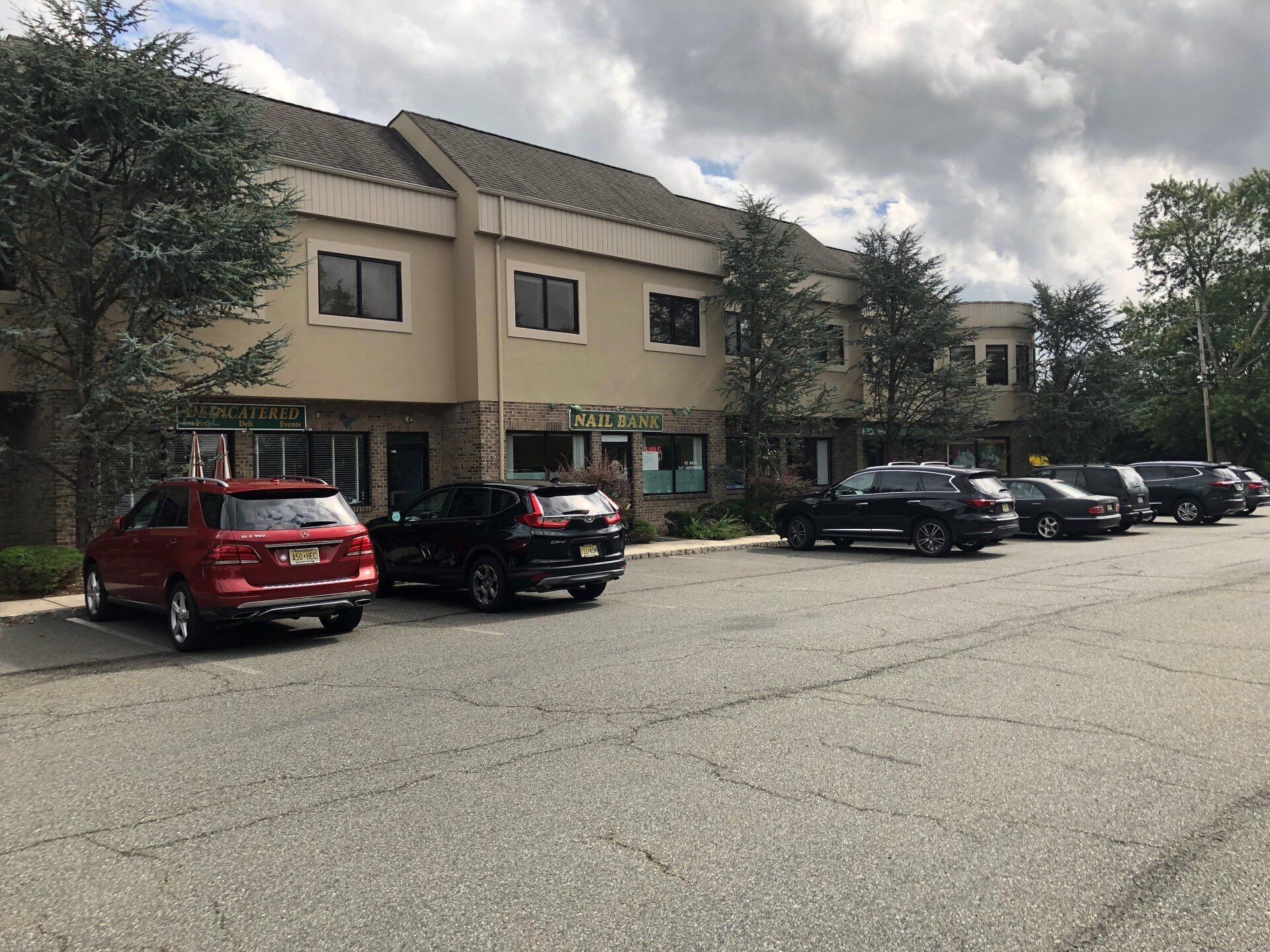 Two-story beige building with several parked cars in front on an asphalt lot; cloudy sky.